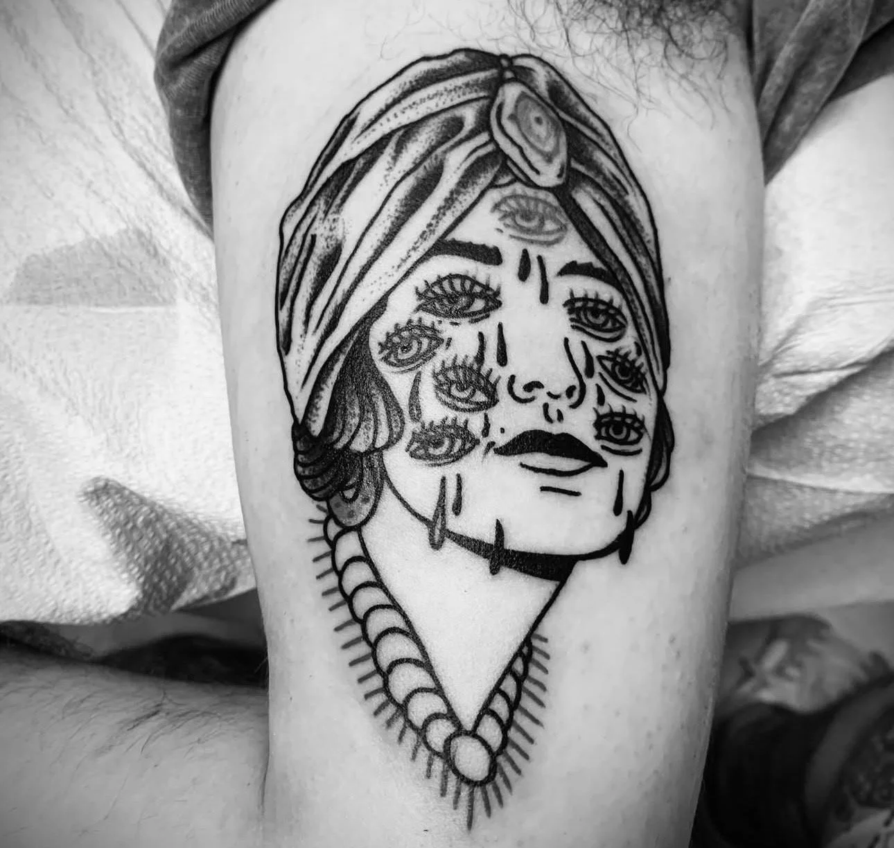 Tattoo of a woman's face with multiple eyes and tears, wearing a headscarf and a necklace.