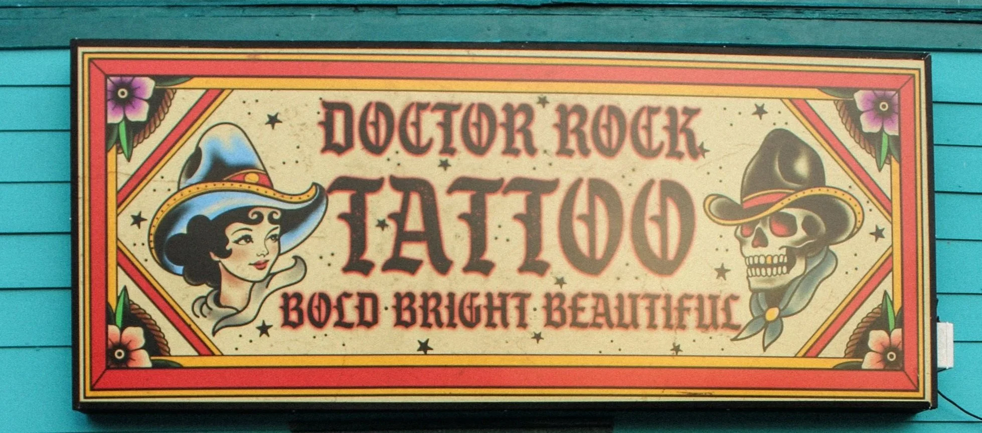 Sign for Doctor Rock Tattoo featuring a woman with a cowboy hat and a skull with a cowboy hat, decorated with flowers and stars.