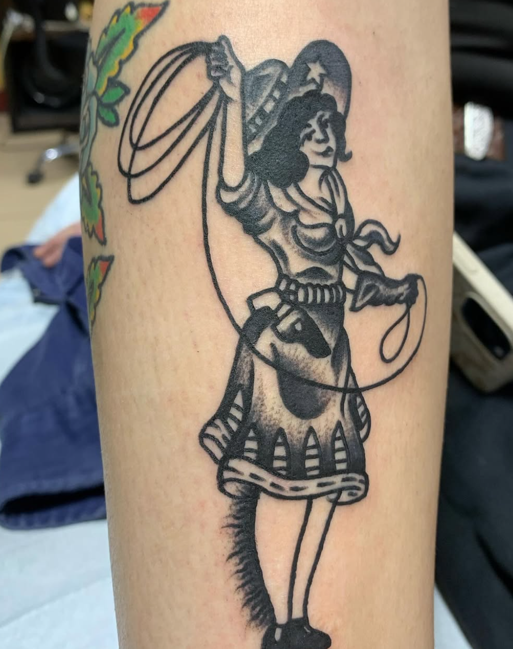 A black and gray tattoo of a young girl dressed as a cowgirl, holding a lasso in one hand and a gun in the other, wearing a hat, bandana, and skirt.