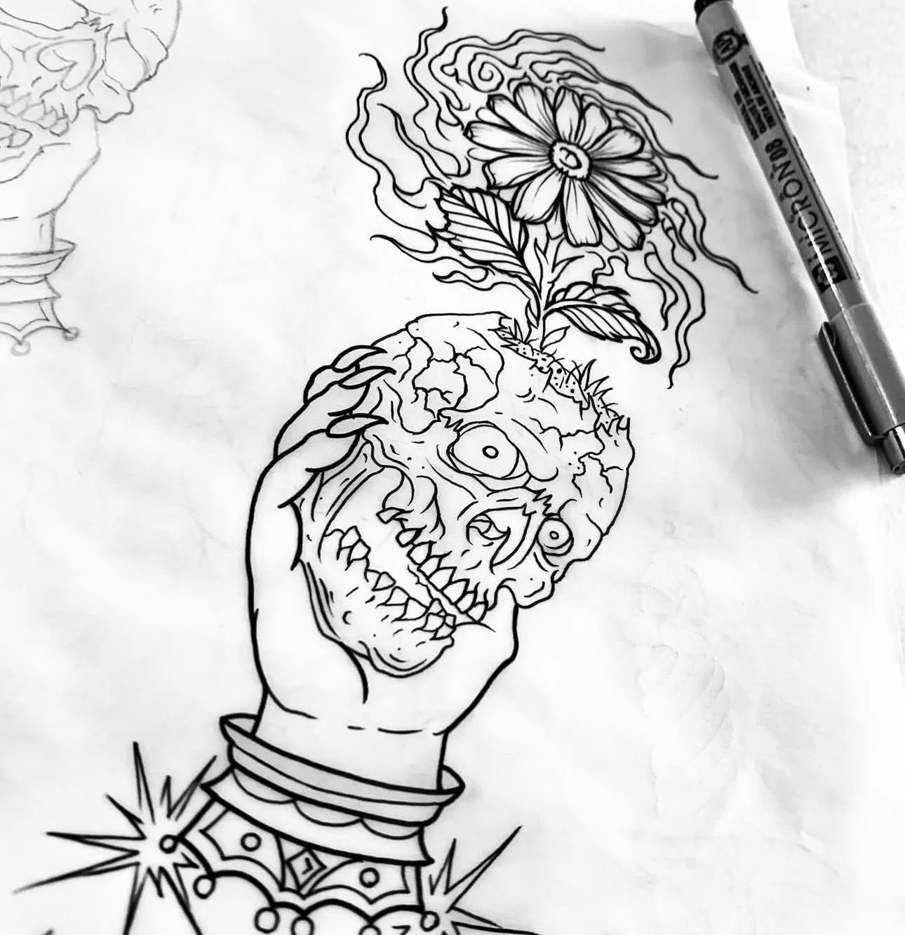 Black and white illustration of a hand holding a skull with a large flower growing out of it, detailed tattoo-style lines, with part of a second design visible in the upper left corner, and a marker pen resting on the paper.