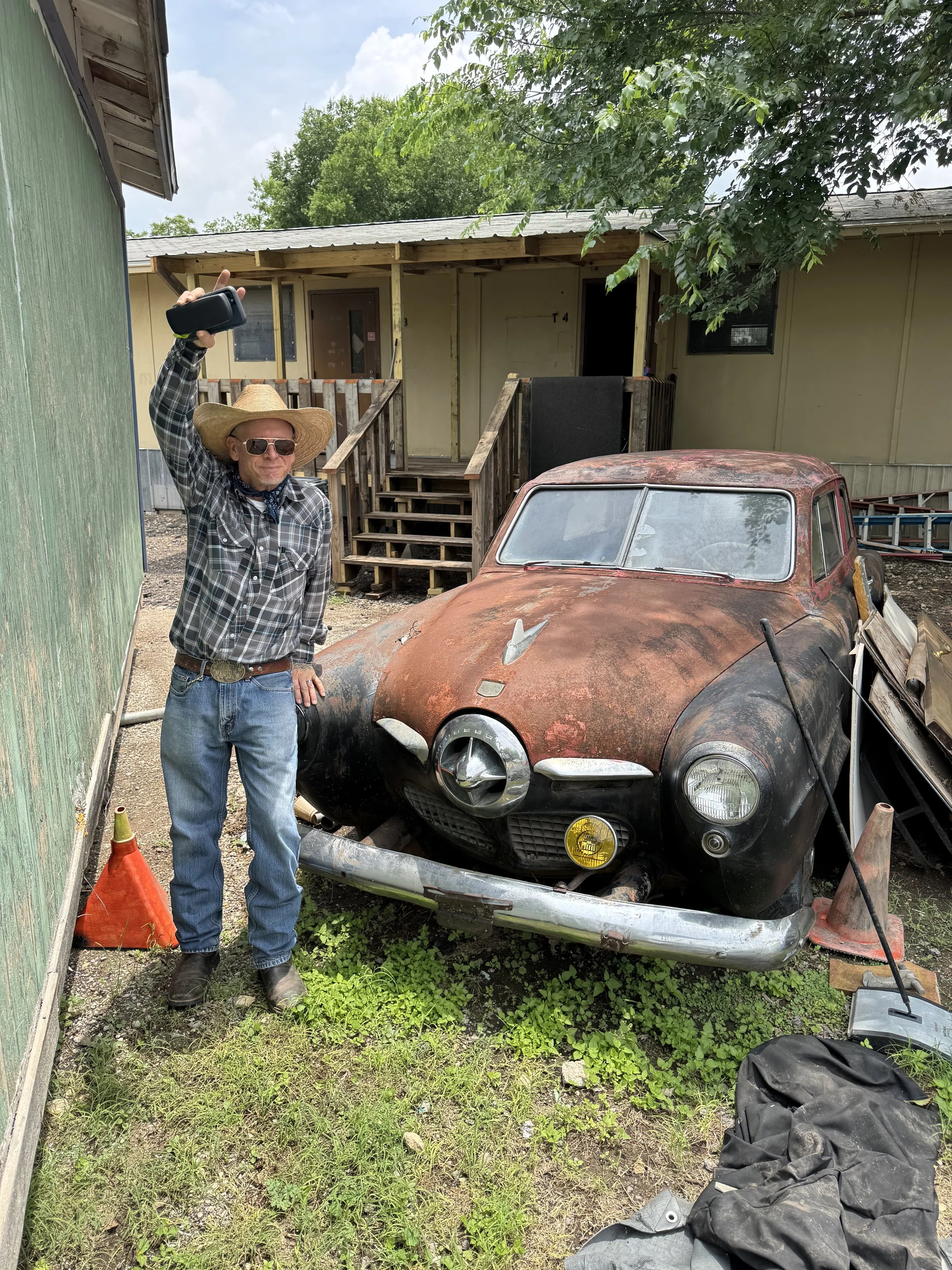 A man wearing a large straw hat, sunglasses, a plaid shirt, and jeans stands next to a rusted, vintage black car with yellow fog lights, in an outdoor area. He is holding a camera or phone in his right hand. There are wooden stairs and doors in the background, along with some greenery and scattered tools or debris on the ground.