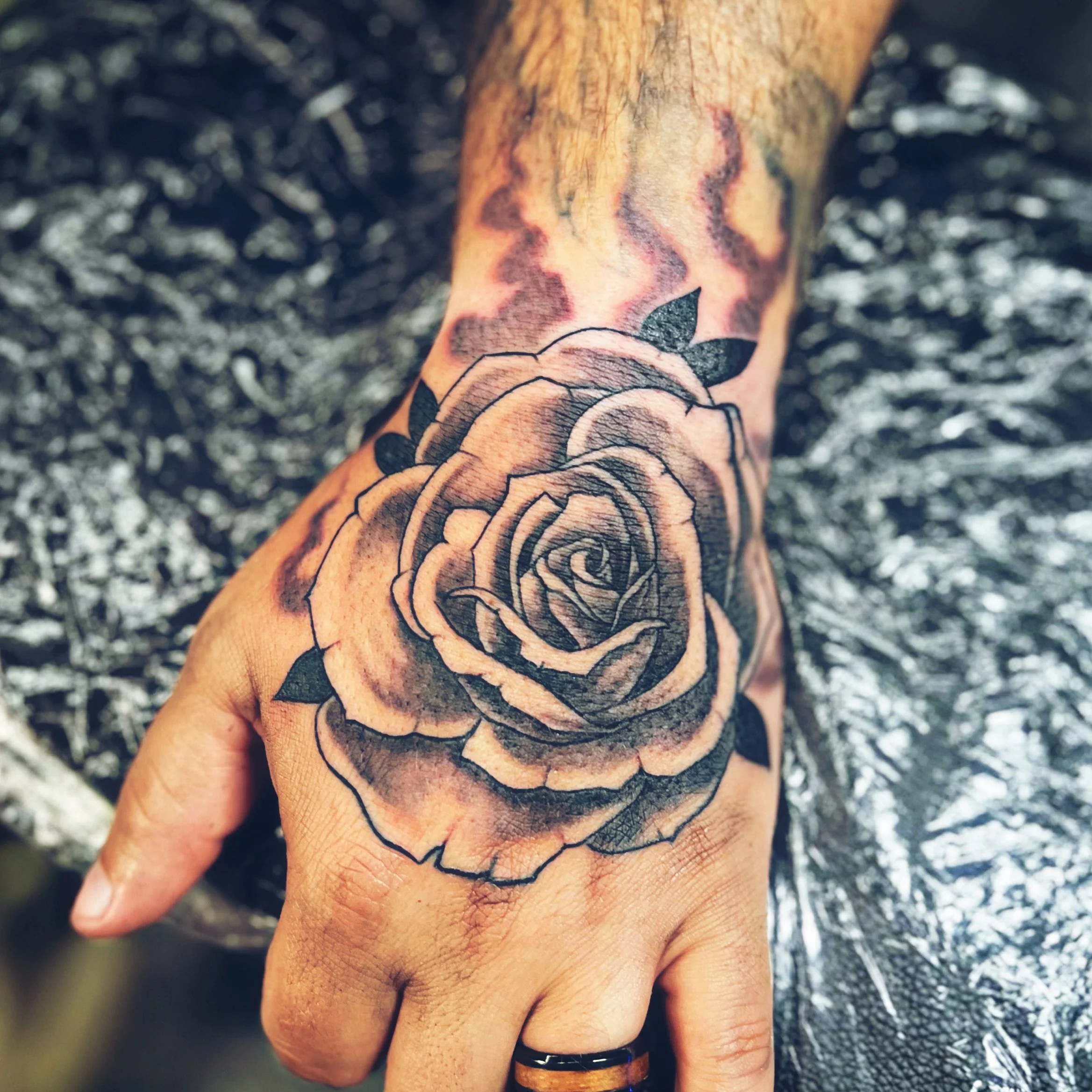 Close-up of a tattooed finger with a rose design on the back of the hand, against a dark, blurry background.