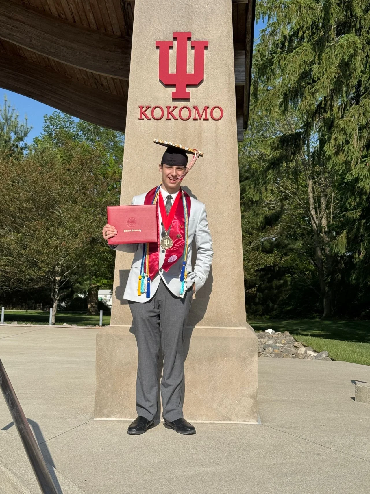 Young man in graduation cap and gown holding a diploma, standing under a large Indiana University sign with trees in the background.