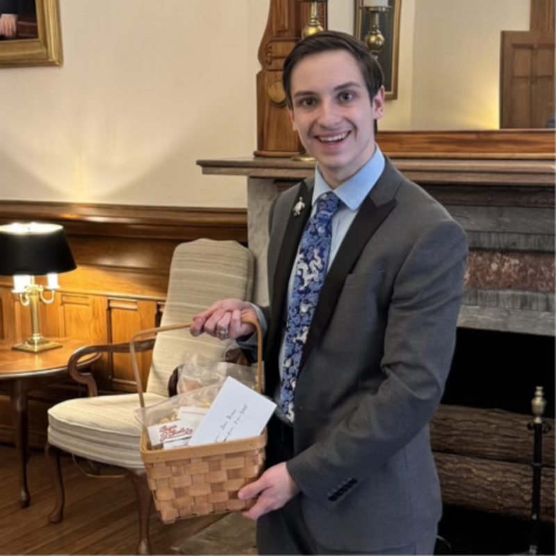 A young man in a gray suit and blue floral tie standing indoors, smiling, holding a small basket filled with popcorn.