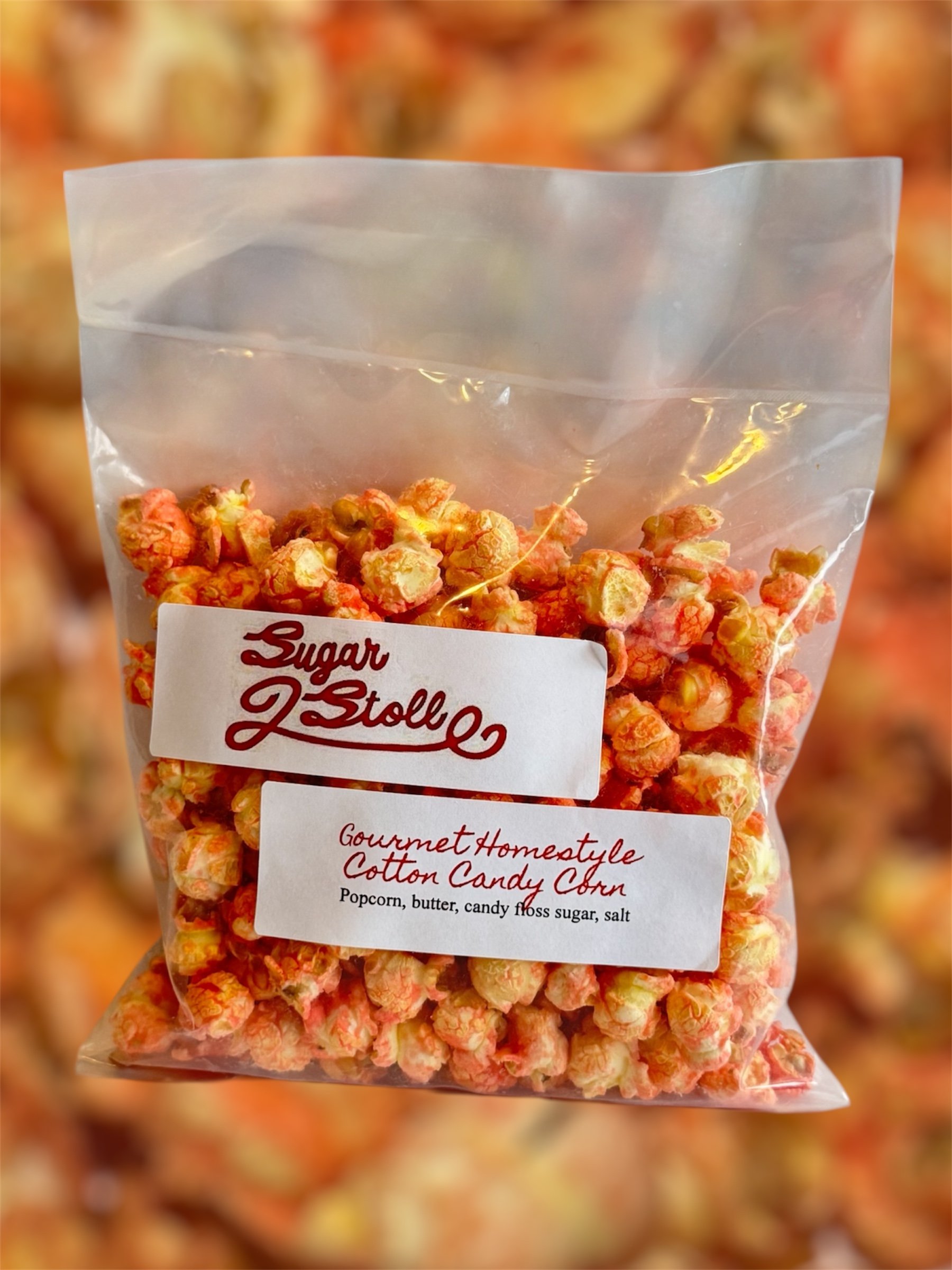 A clear plastic bag of popcorn with a label that reads 'Sugar & Jollies,' describing it as gourmet homestyle cotton candy popcorn with ingredients including popcorn, butter, candy floss sugar, and salt. The background is blurred with similar popcorn bags.