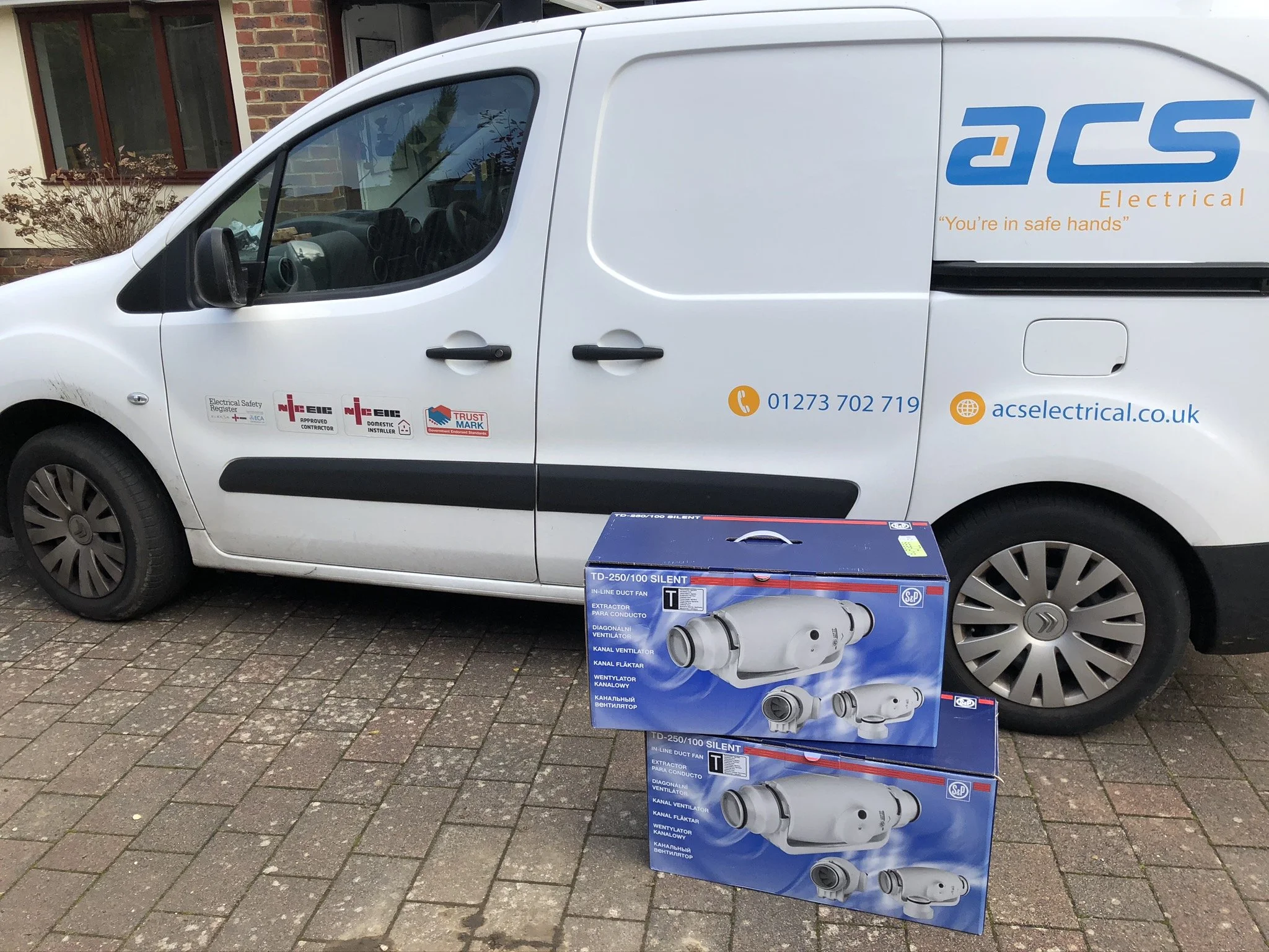 White van with company logo and contact information parked in front of a residential building, with two boxed ventilation fans on the ground in front of it.