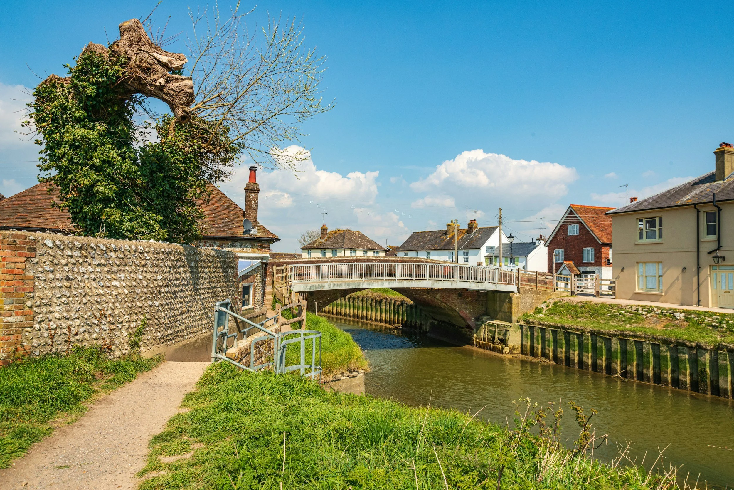 A small footbridge over a canal in a village, with old brick and plaster houses on either side, under a bright blue sky with scattered white clouds.