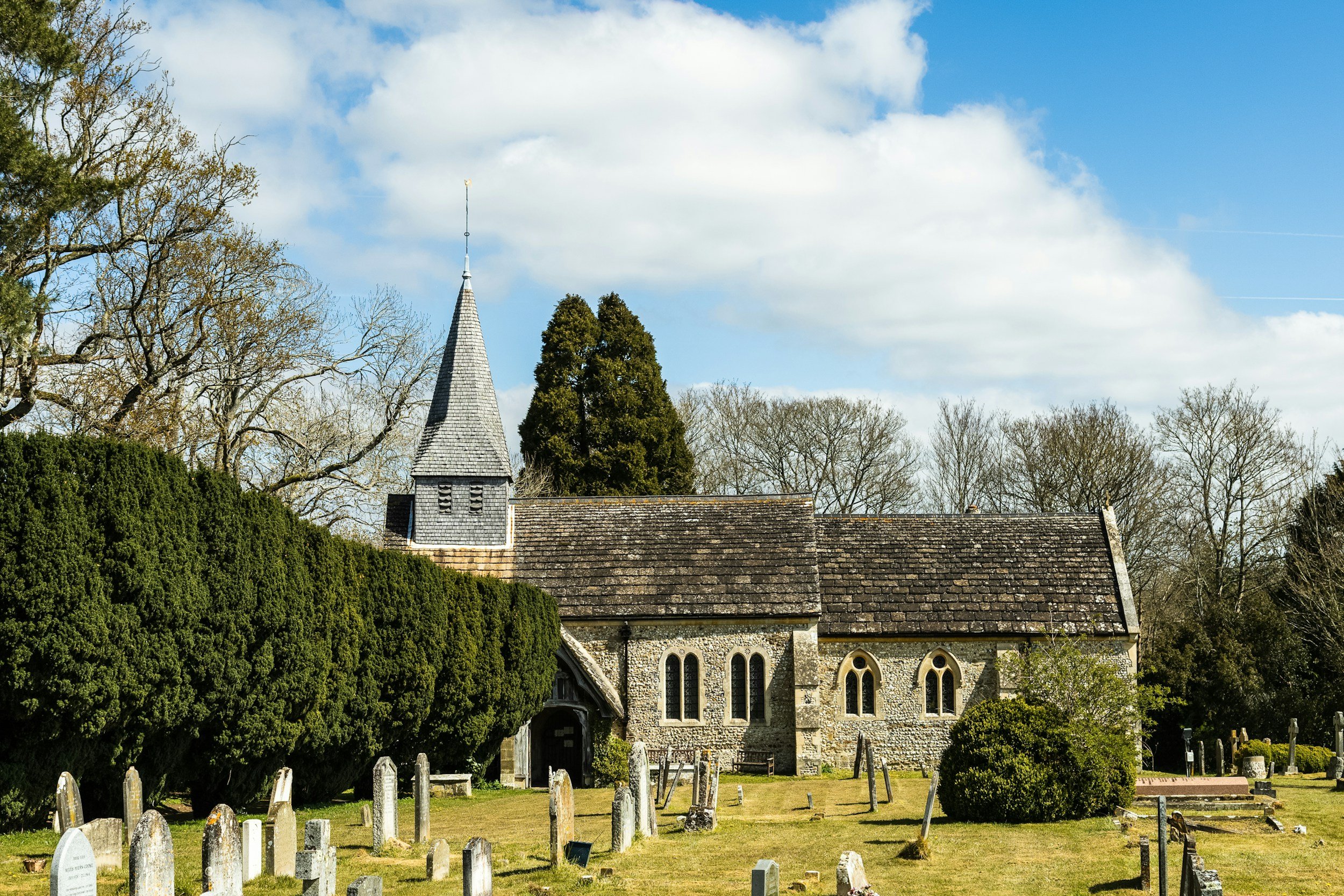 View of a small church with a steeple, surrounded by trees and a graveyard with tombstones, under a partly cloudy sky.