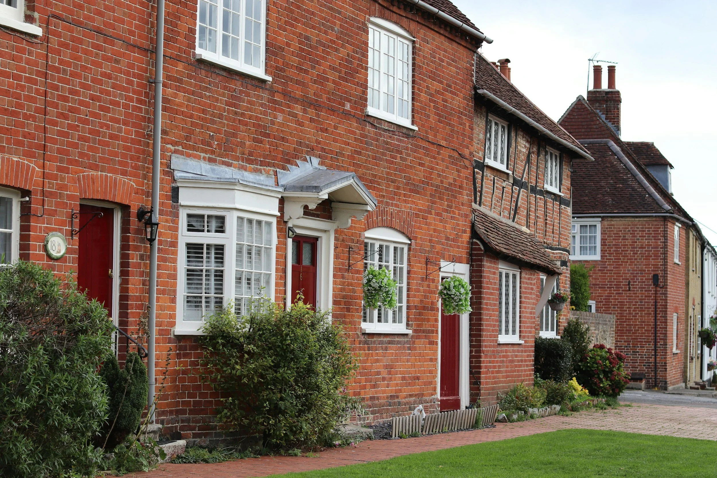 A row of brick houses with white-framed windows and red doors, front gardens with bushes and hanging flower baskets, and a brick sidewalk in front.