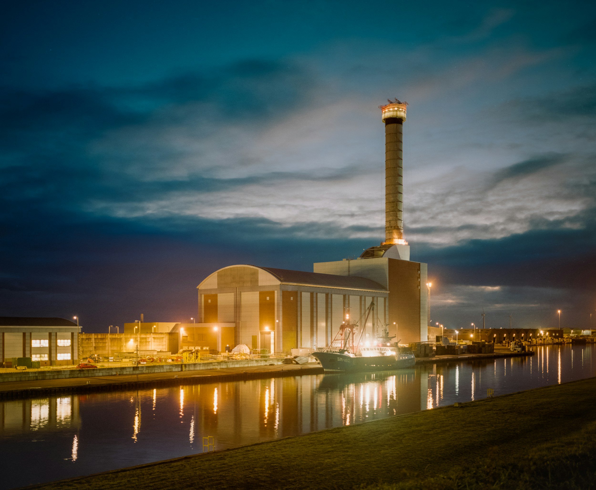Night view of an industrial facility near water, with a tall smokestack illuminated at the top. The water reflects the lights and structures, and the sky is dark with some cloud cover.