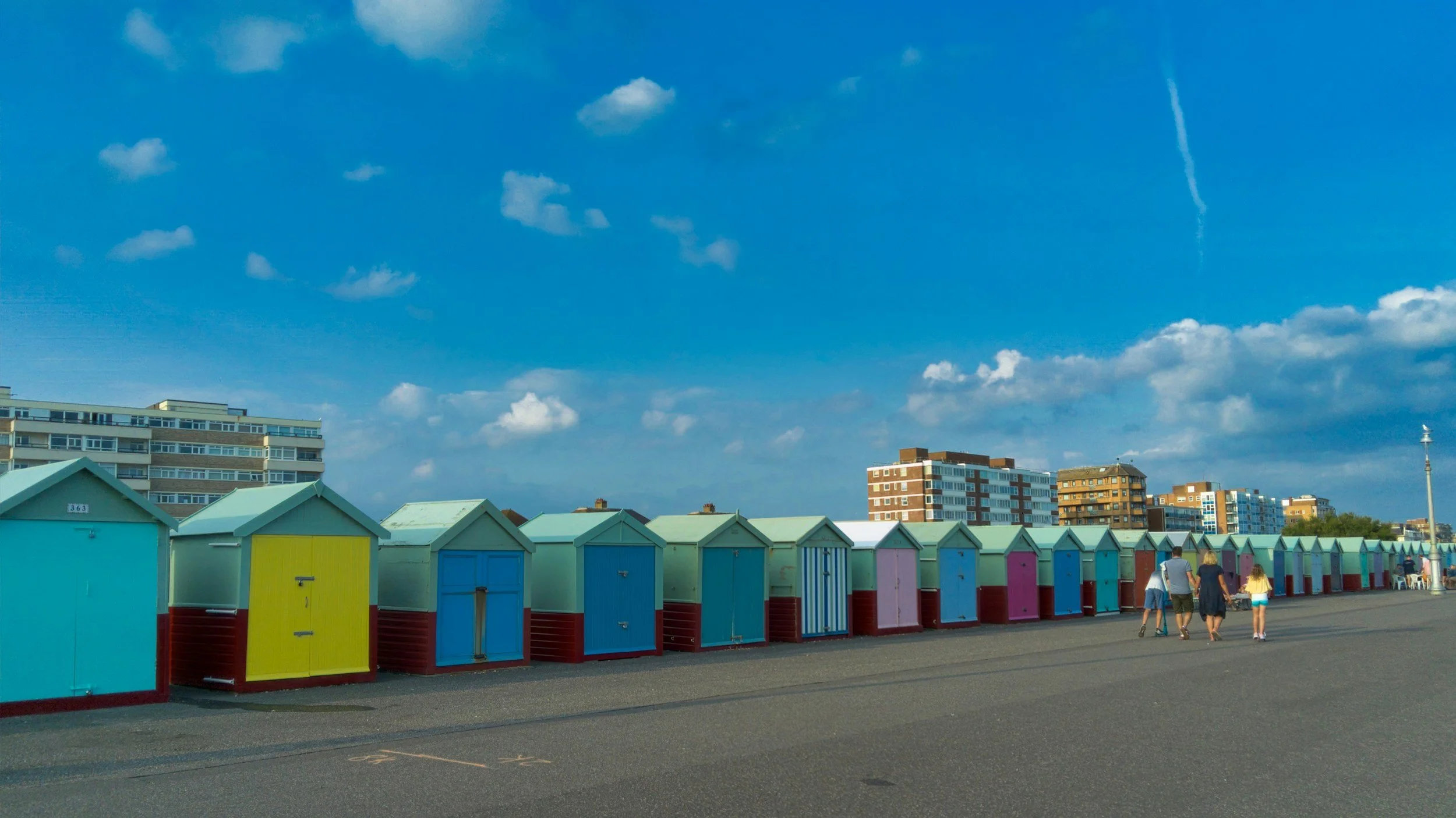 Colorful beach huts lined up along the boardwalk with a few people walking, under a blue sky with scattered clouds and high-rise buildings in the background.