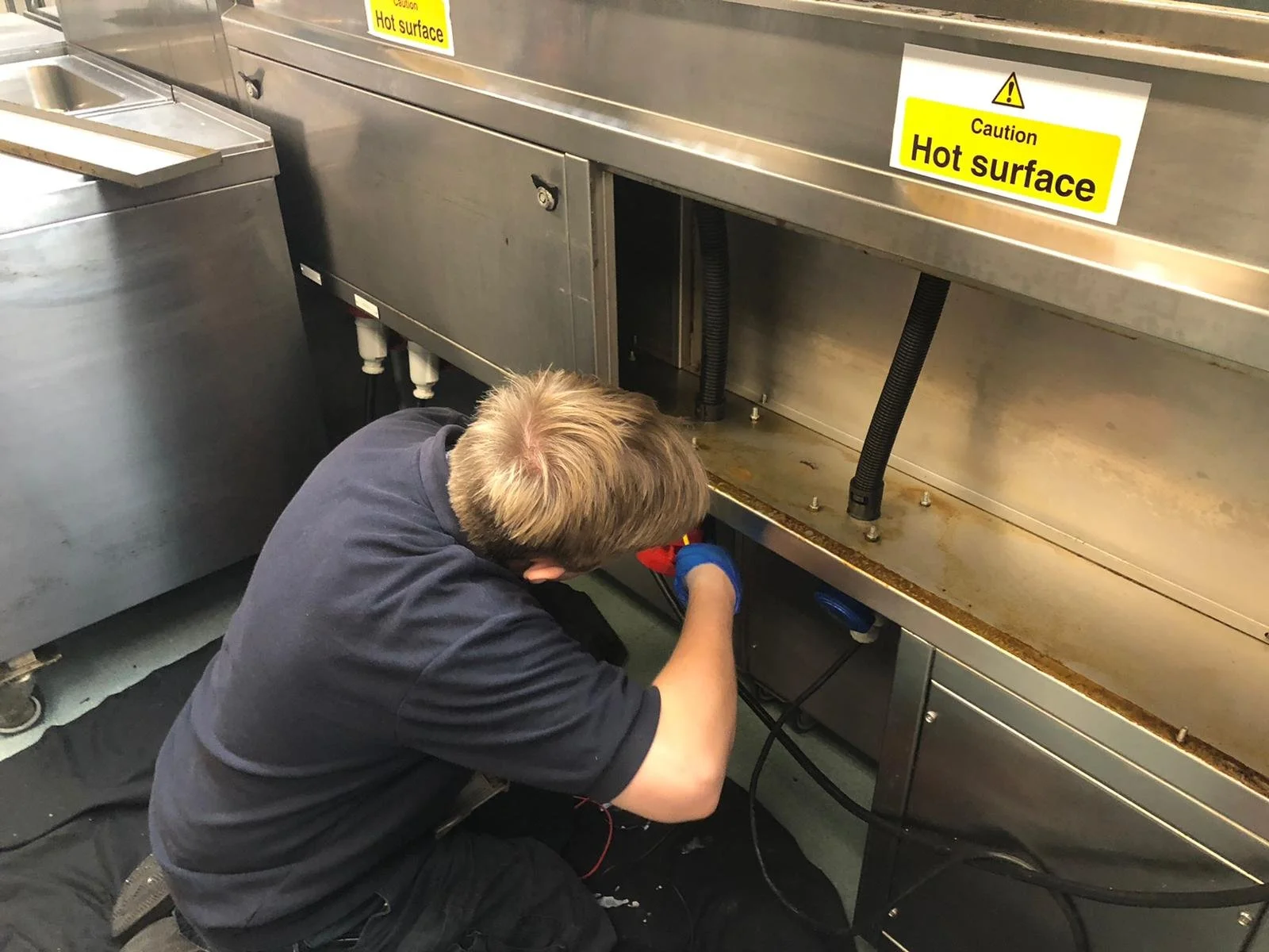 A person working under a stainless steel kitchen appliance marked with a yellow and black