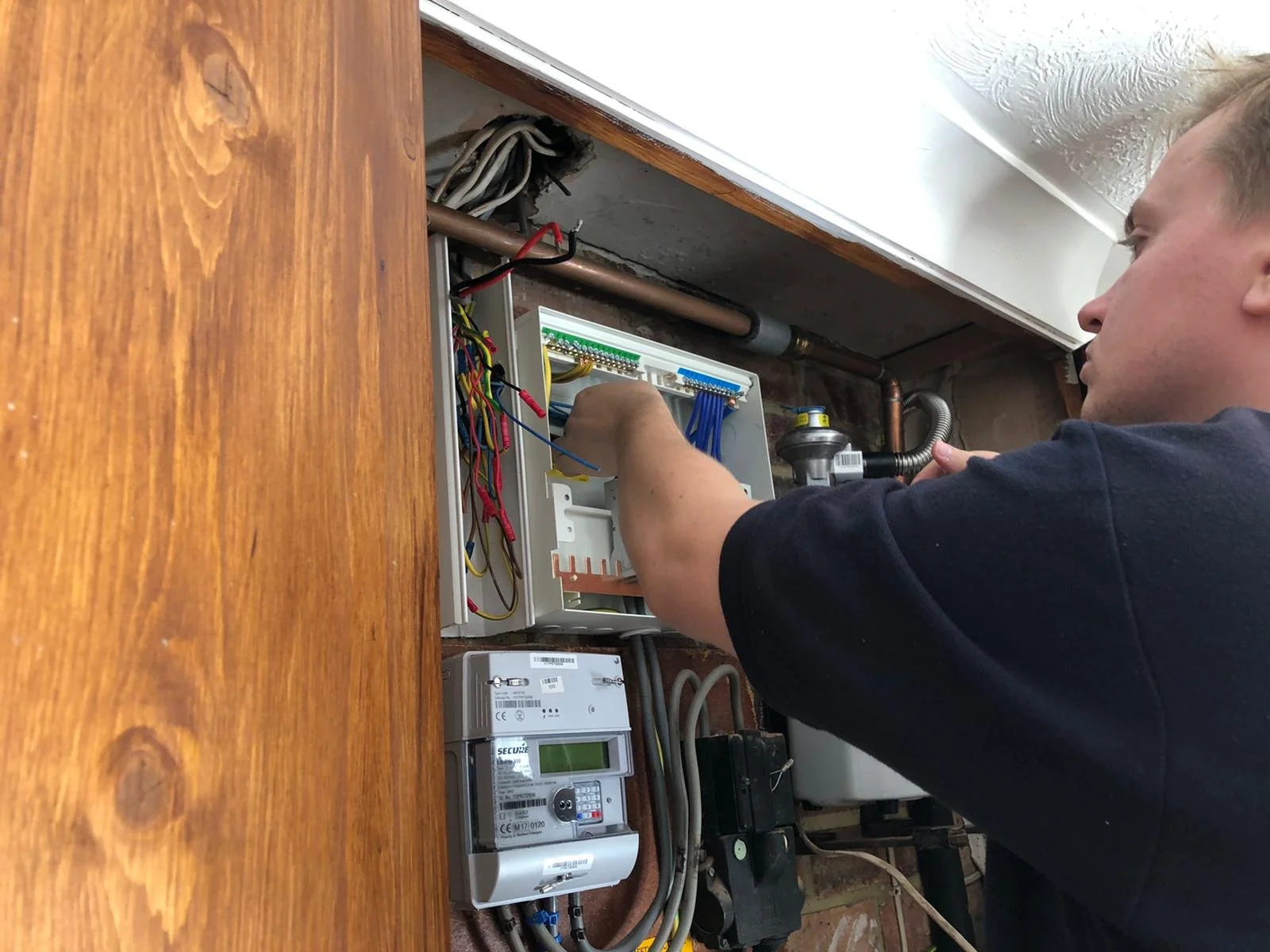 A man working on an electrical panel, installing or repairing wires in a utility box on a wall, with various electrical components around.