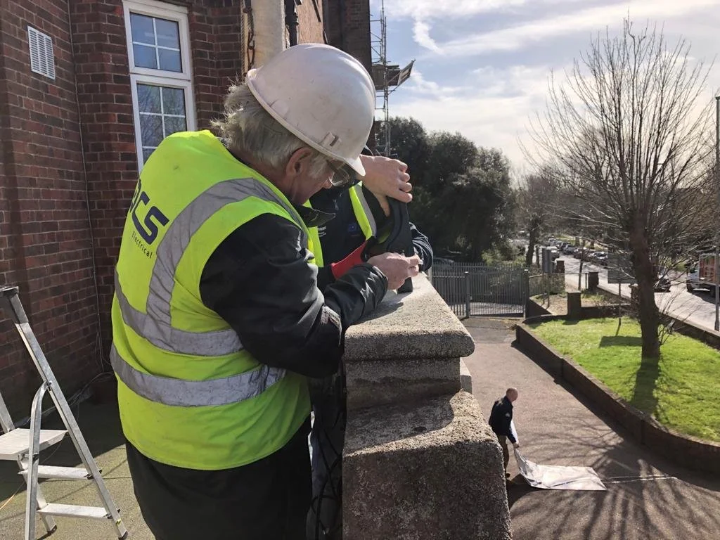 Construction worker in a yellow safety vest, white helmet, and safety glasses working on a building ledge, with another worker below handling a large sheet in an outdoor setting with trees and a sidewalk.