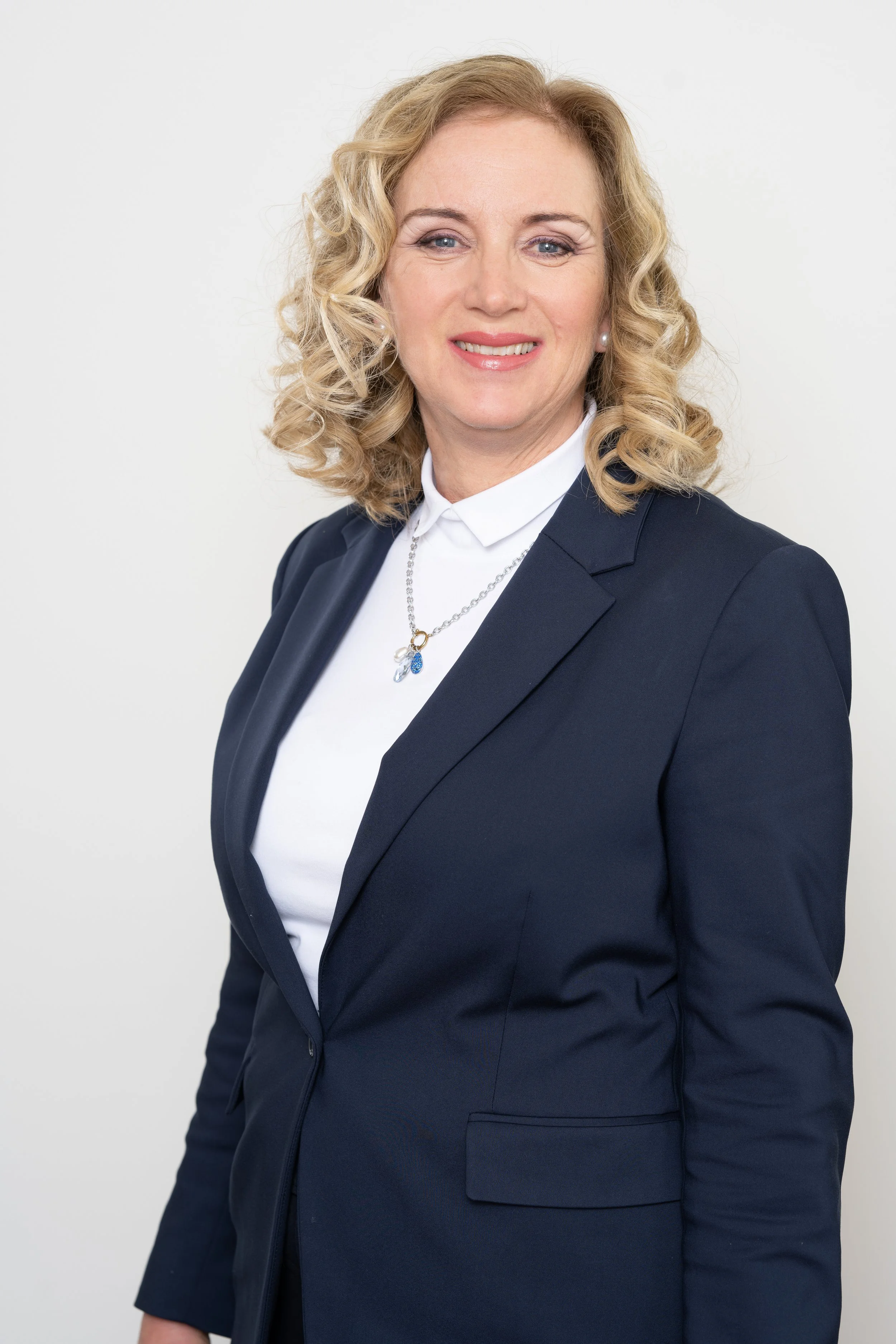Porträt einer lachenden Frau mit blonden, gelockten Haaren, in dunklem Blazer und weißem Hemd, vor weißem Hintergrund.
