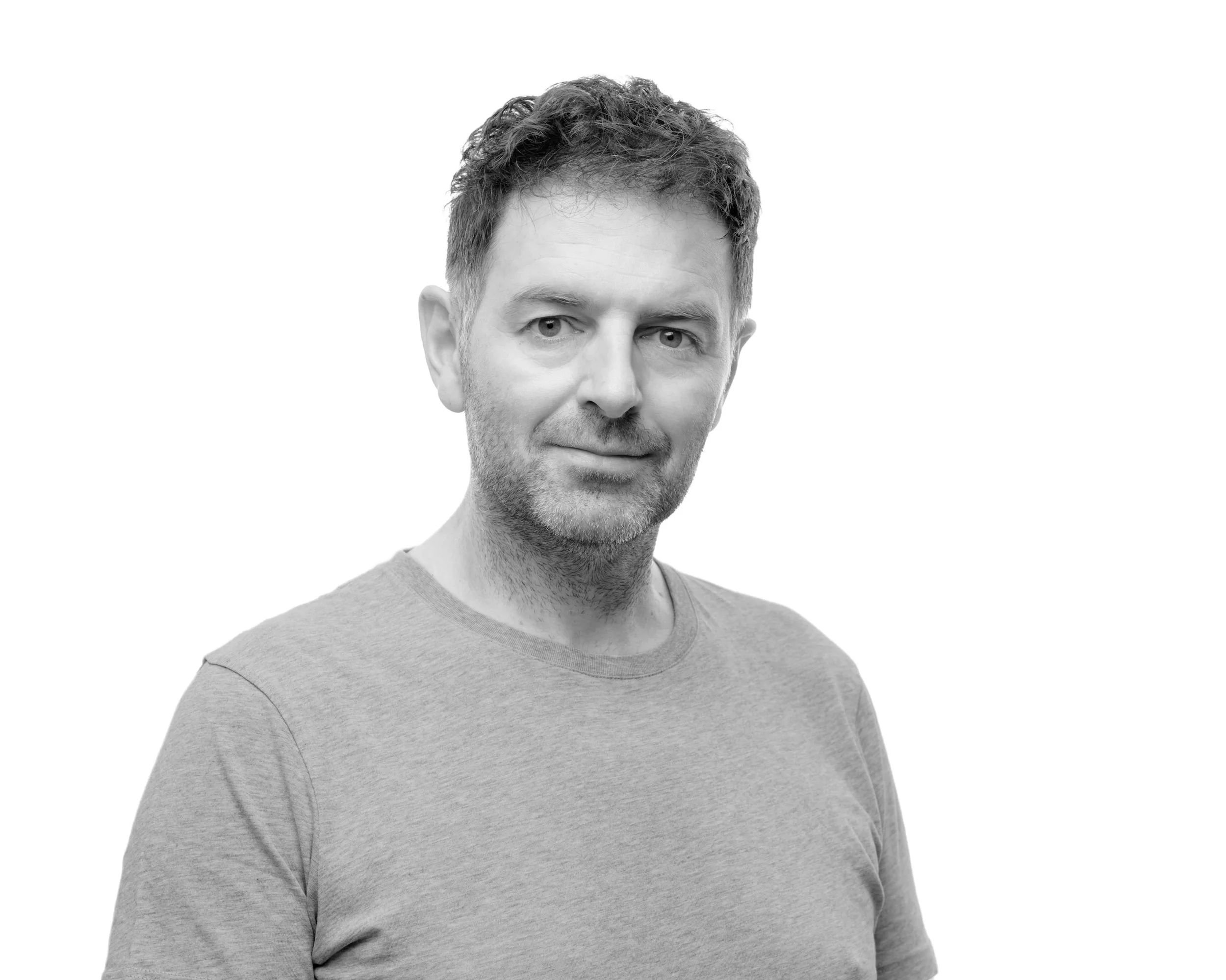 Black and white portrait of a man with short, wavy hair, slight beard, wearing a plain t-shirt, looking into the camera with a neutral expression.