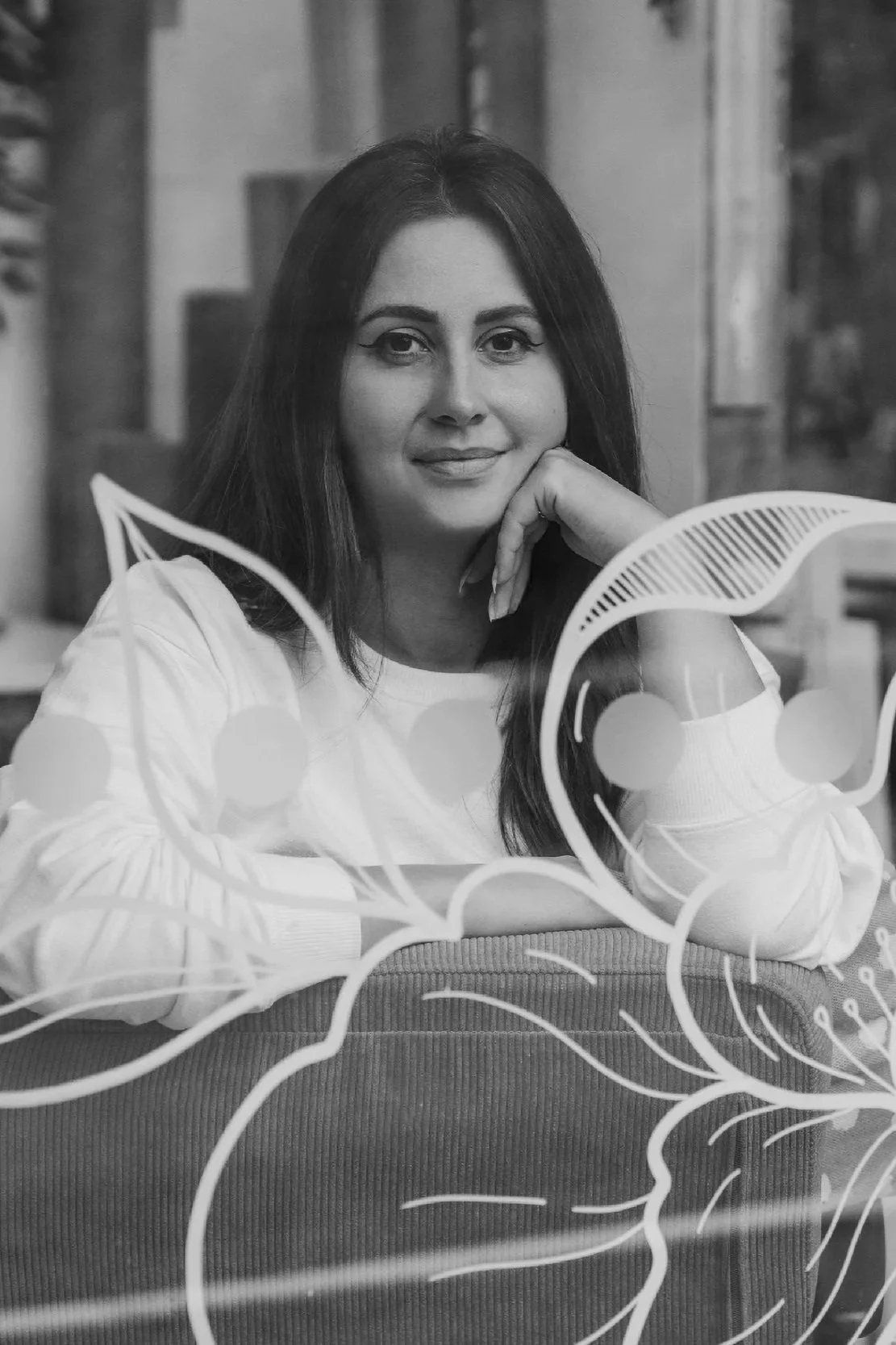 A woman with dark hair, wearing a white top, sitting at a table with her chin resting on her hand, looking at the camera. There is a decorative art piece with human profiles and abstract lines in the foreground, and a blurred background.