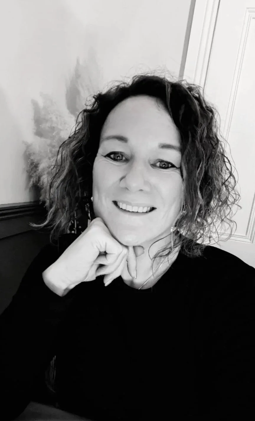 Black and white photo of smiling woman with curly hair, resting her chin on her hand, indoors.