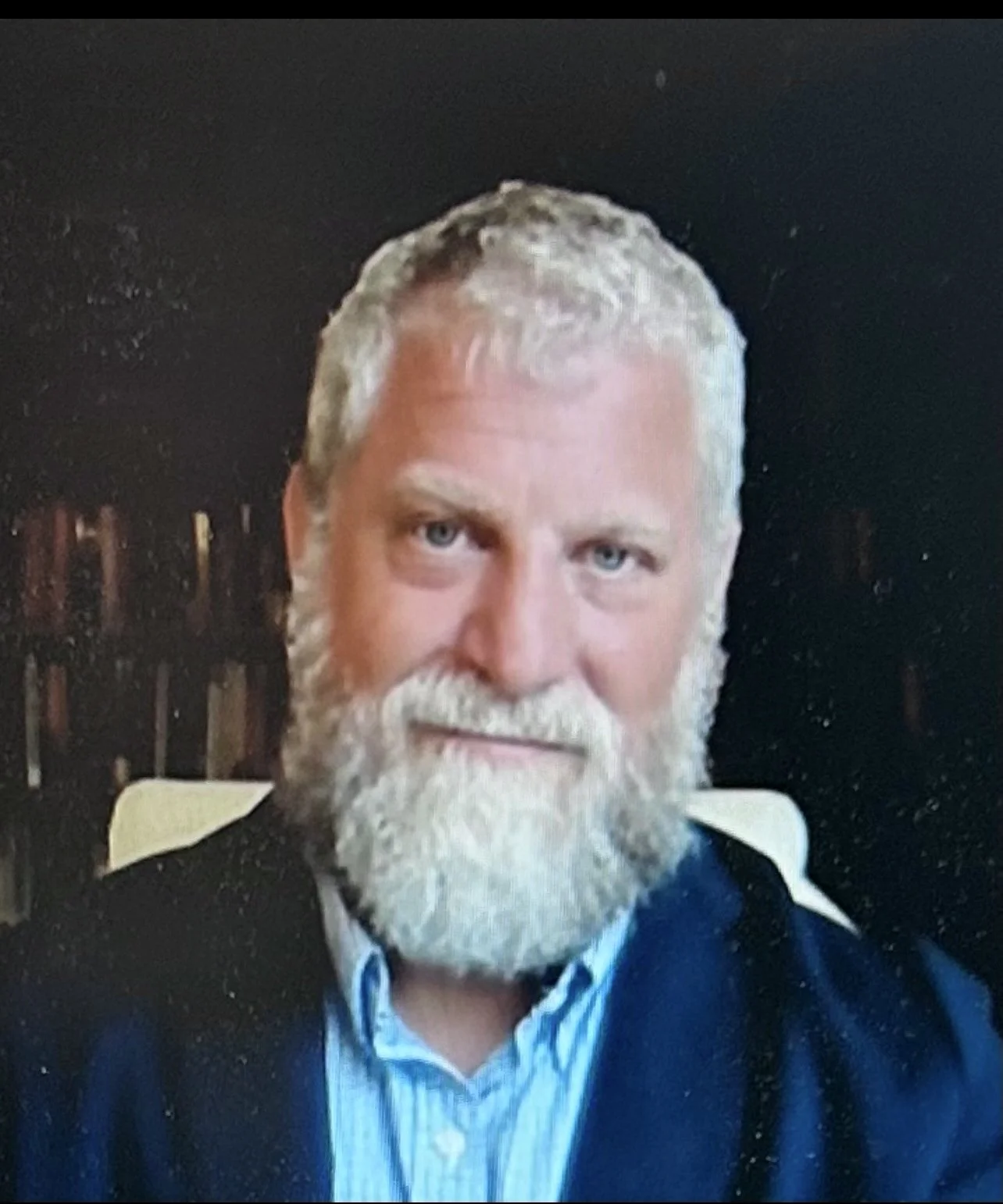 A man with a full white beard and gray hair, wearing a dark jacket and light collared shirt, sitting in a room with bookshelves in the background.