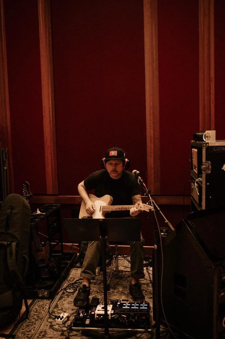 A man wearing headphones and a black cap playing an acoustic guitar in a recording studio.