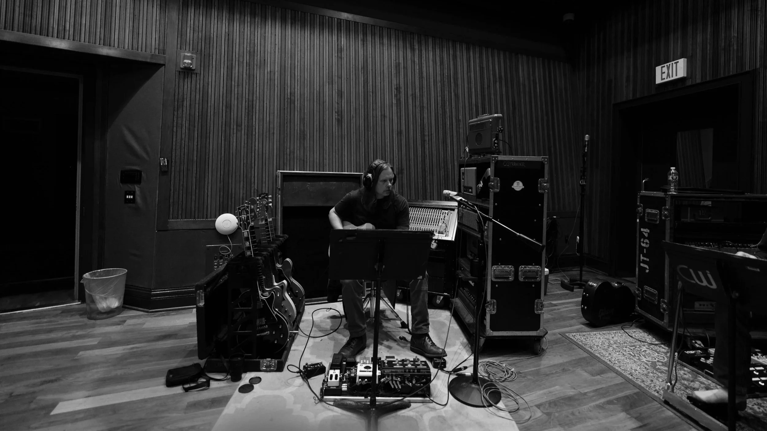 Justin Ostrander in a Nashville Studio wearing headphones sitting at a sound mixing console in a recording studio. Multiple guitars, equipment cases, and audio gear surround her, with wood-paneled walls and an exit sign visible in the background.