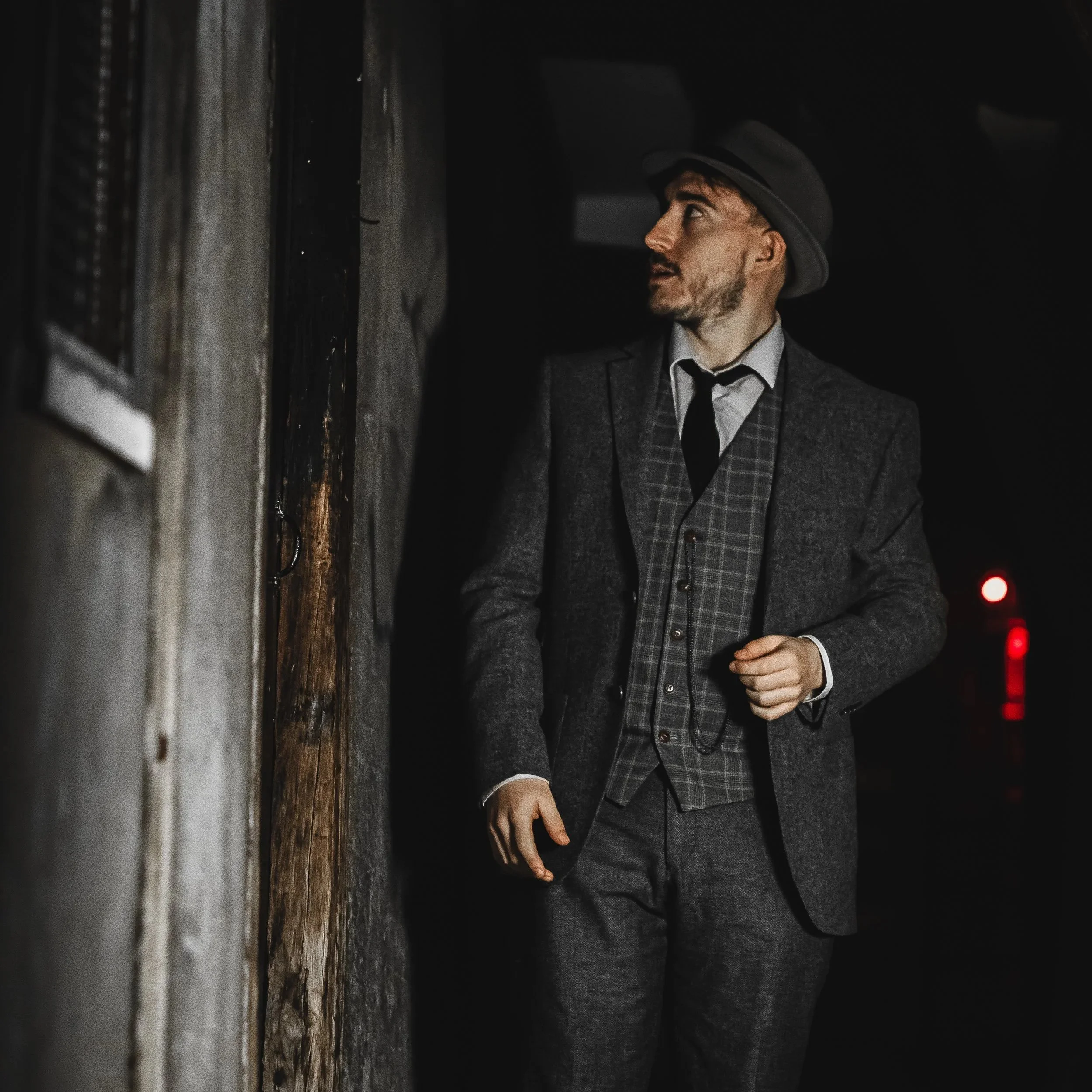 A man dressed in a gray chequered suit, white shirt, and black tie, wearing a gray fedora hat, standing and looking upward in the Shambles.