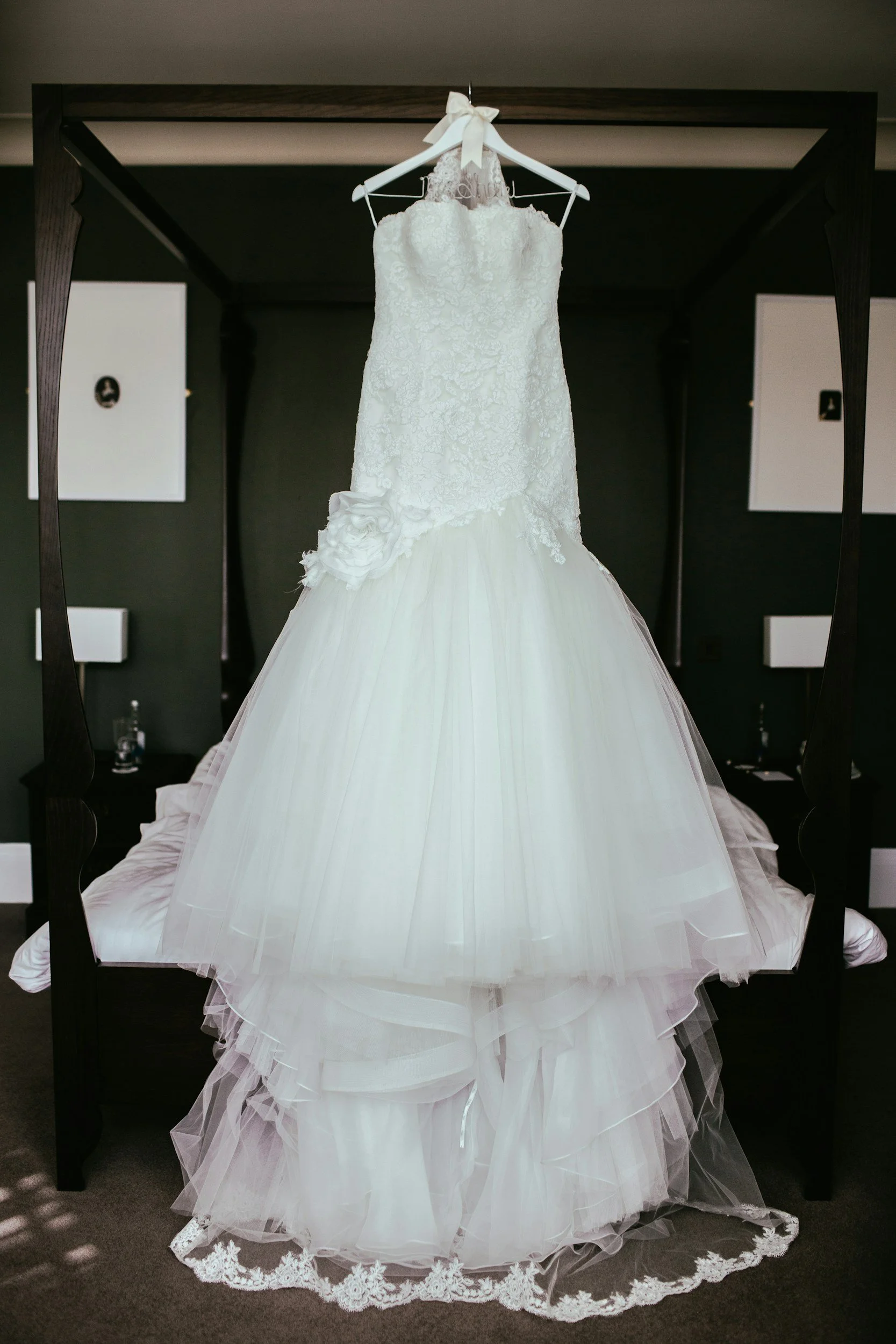 A white wedding gown hanging on a hanger with a bow, displayed on a black furniture piece, with a layered tulle skirt with lace trim at the bottom, in a room with dark walls and framed pictures.