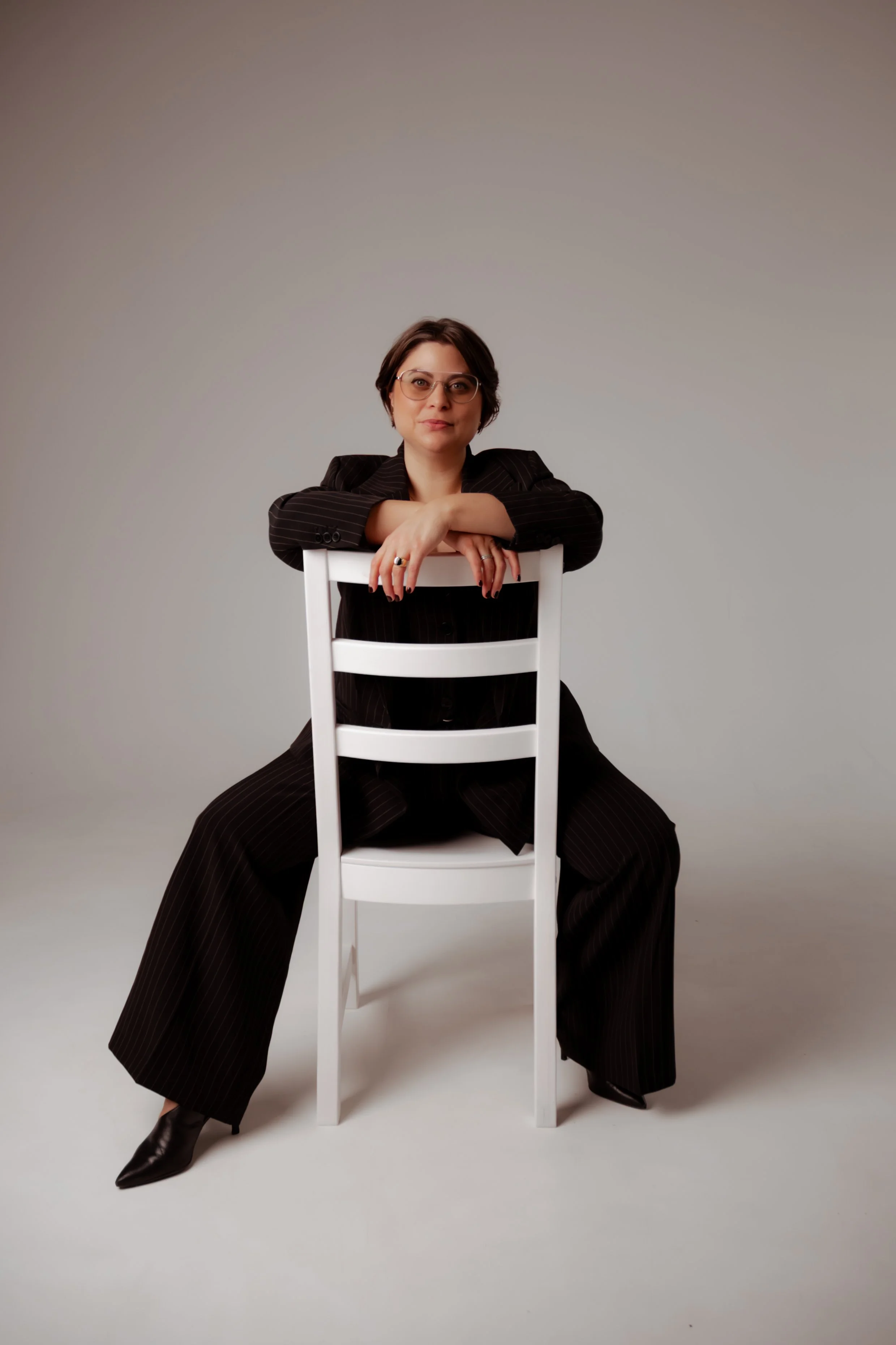 A woman with short dark hair and glasses sitting on a white chair against a plain light grey background. She is wearing a black pinstripe suit, black boots, and has her arms resting on the back of a white chair in front of her. She is looking directly at the camera.