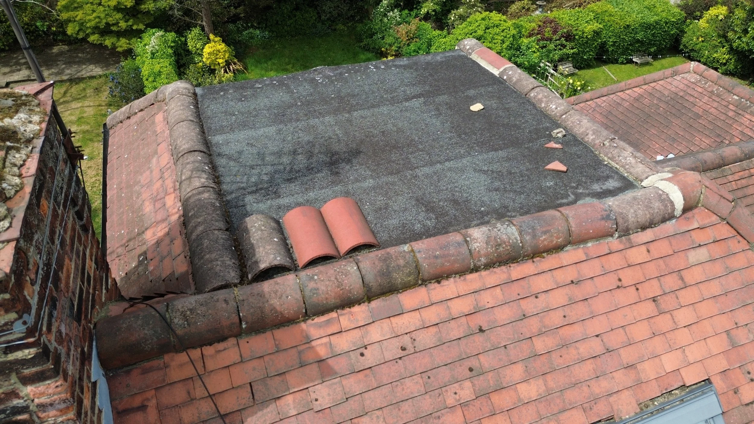 A rooftop with red clay tiles and a black rubber covering, with several broken roof tiles and some clay tiles rolled up at the edge, overlooking a green garden with trees and benches.