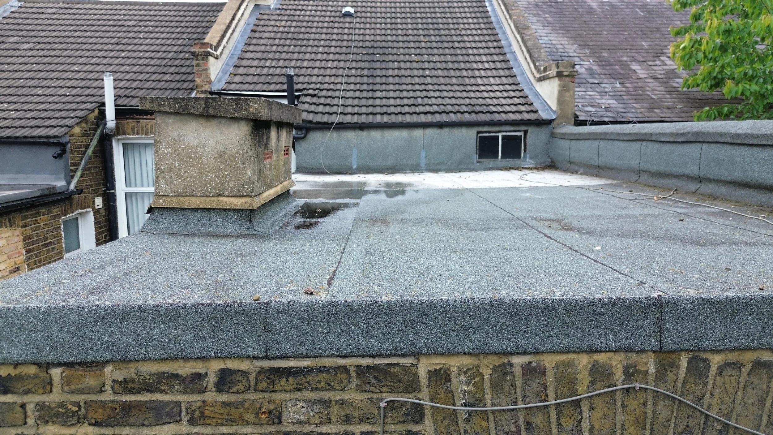 View of rooftops with tiled and flat roofing, chimneys, and a small window, with a branch of a tree on the right side.