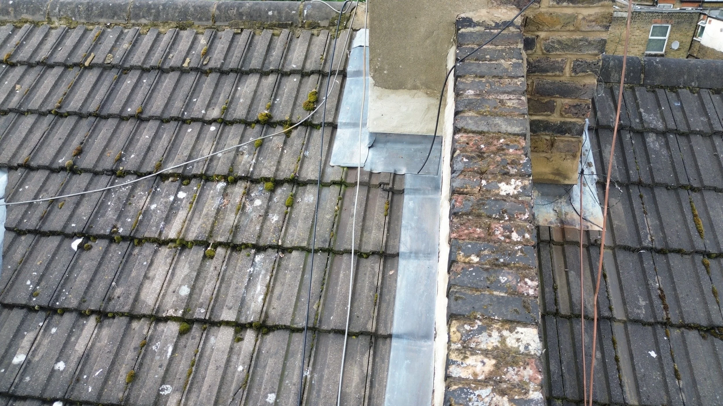 A close-up view of a rooftop with weathered, moss-covered tiles, metal flashing, and a brick chimney, with rooftops and buildings visible in the background.