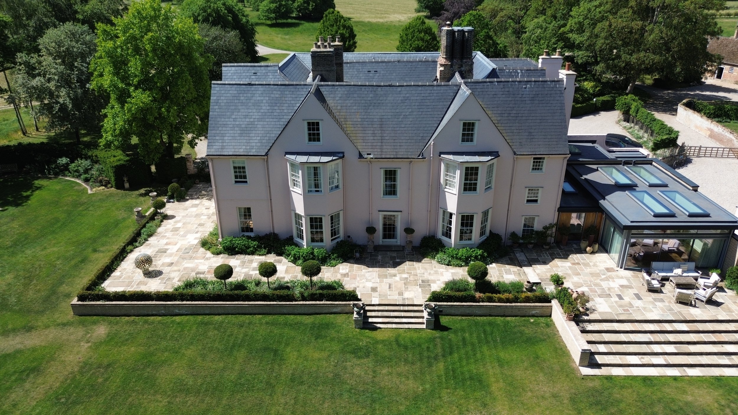 An aerial view of a large, elegant house with a gray roof, white exterior walls, and multiple chimneys. The house is surrounded by a well-maintained lawn and a large patio area with outdoor furniture, and there are trees and greenery in the background.