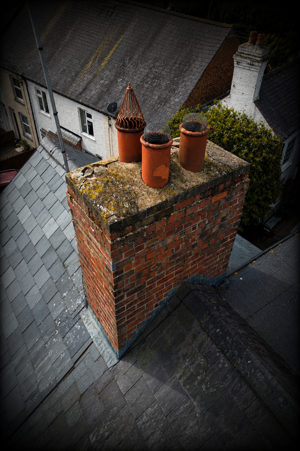 A rooftop chimney with three terracotta pots on top, one of which has a metal mesh cover. The chimney is made of red bricks with some moss and dirt on the top. Surrounding rooftops and a variety of trees are visible in the background.