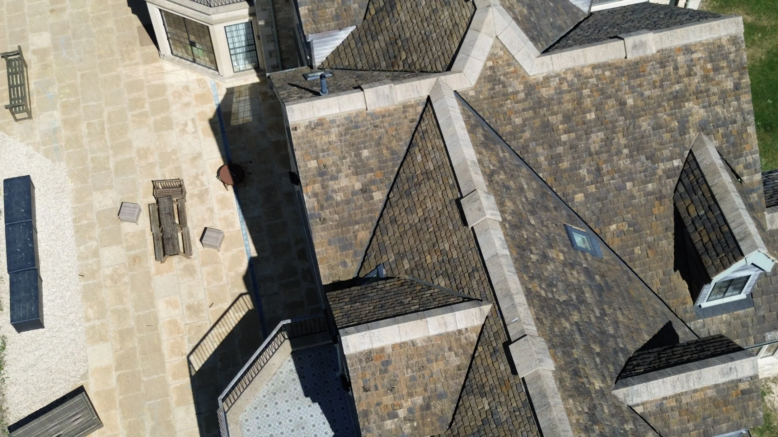 An aerial view of a residential building with a brown shingle roof and a patio area with wooden tables and chairs, surrounded by a gravel and stone paved yard.