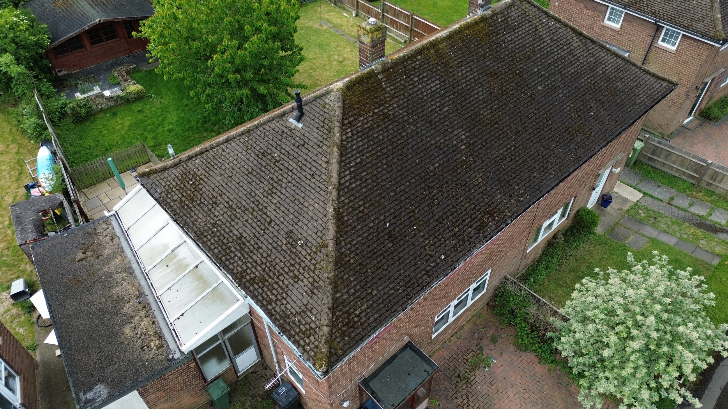 An aerial view of a brick house with a sloped tiled roof, surrounded by a backyard with green grass, trees, and a patio area with outdoor furniture and a small swimming pool.