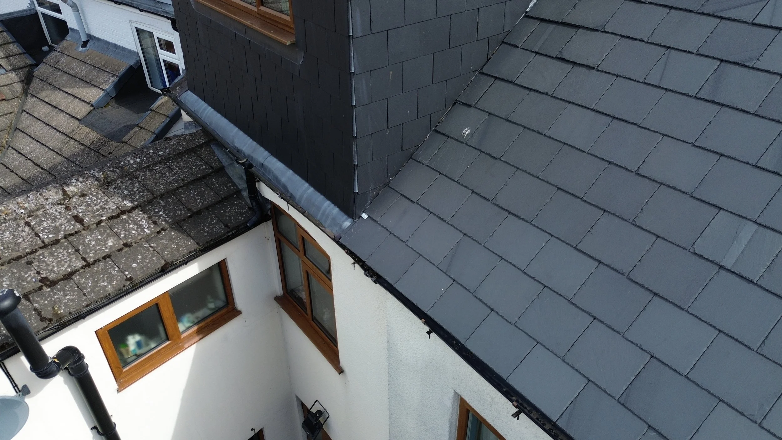 Aerial view of rooftops on a building with different types of tiles, including plain slate and moss-covered tiles, and a black downspout on a white wall. The building has large windows with brown frames.