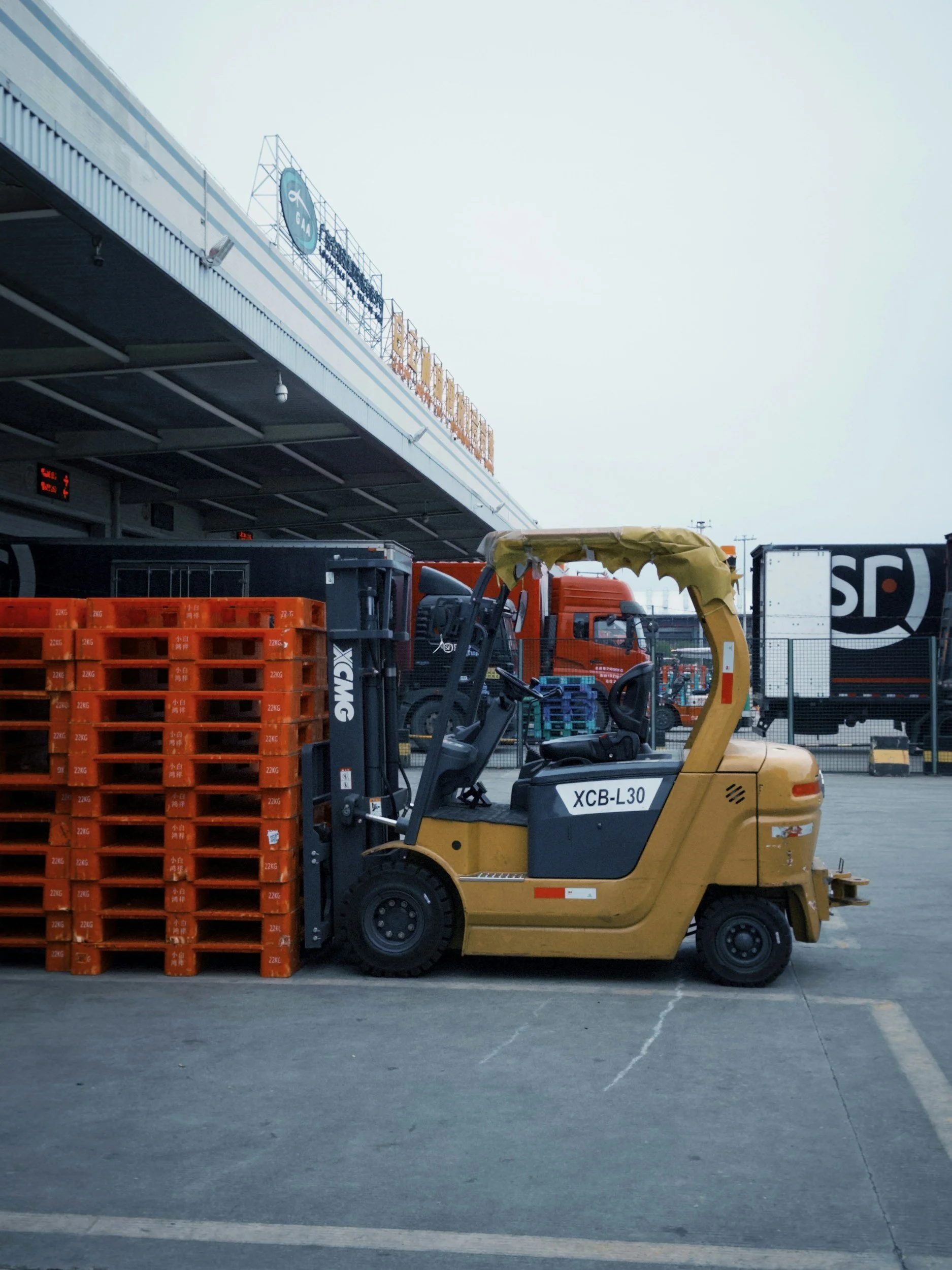 A yellow forklift truck moving orange plastic crates near a warehouse loading dock with signage on the building and a cloudy sky overhead.