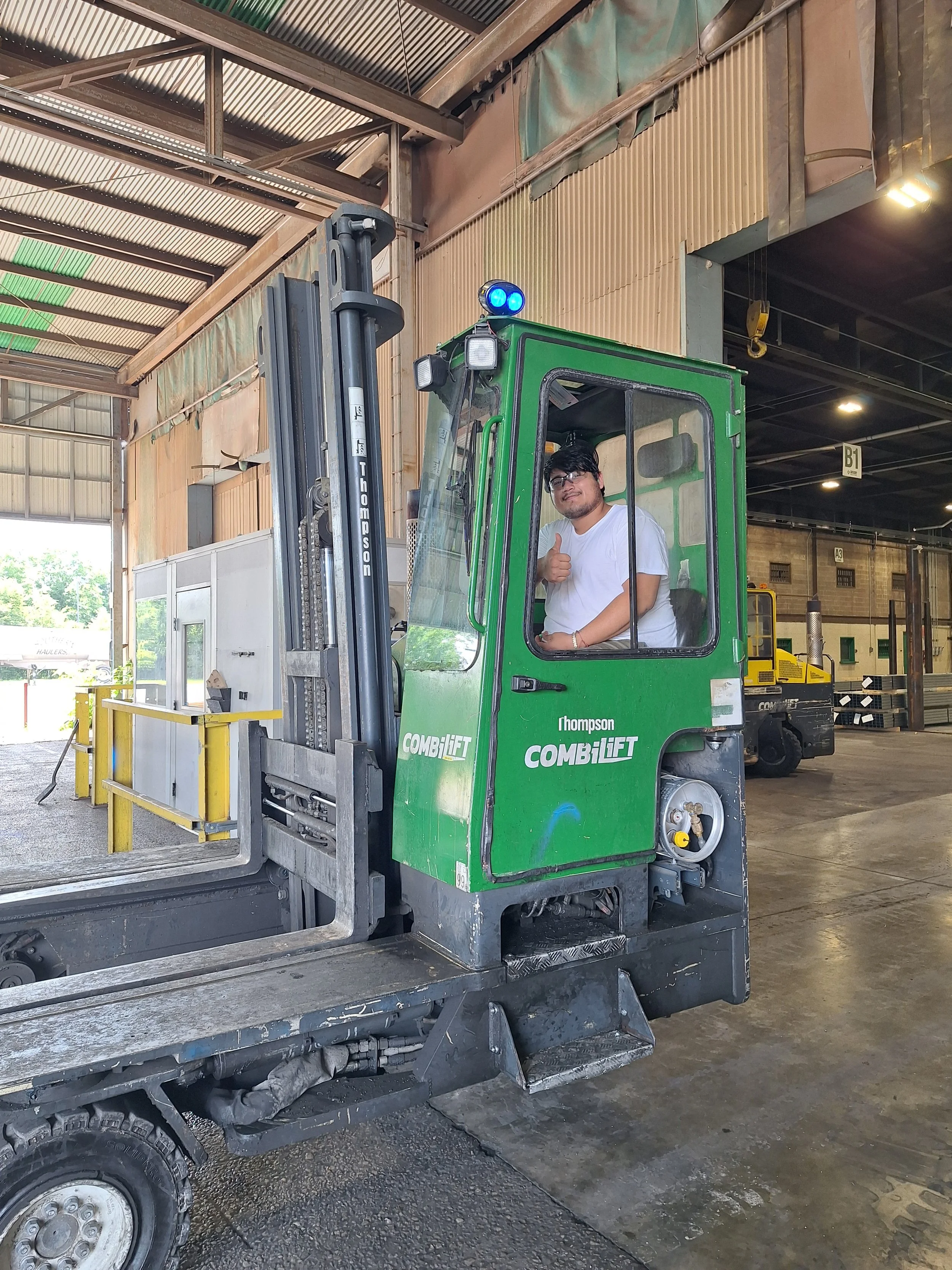 A man is sitting inside the cab of a green forklift with his thumb up, in a warehouse or industrial facility.