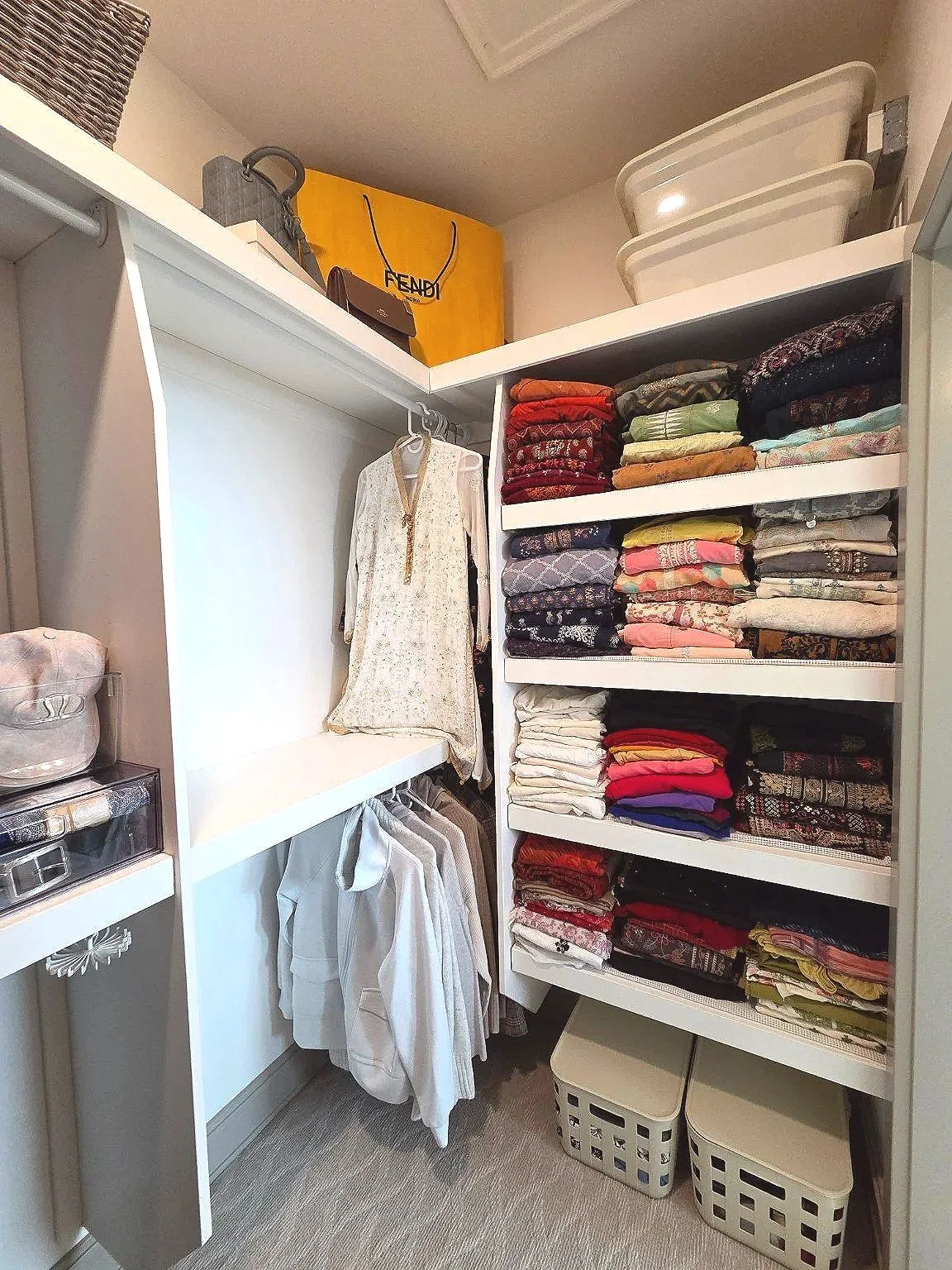 A walk-in closet with organized shelves of folded clothes on the right, hanging garments on the left, and storage boxes on the top shelf. Professional home organization in Saskatoon and area.