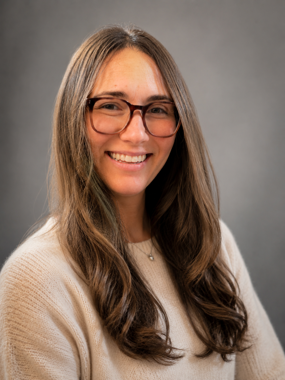 A woman with long brown hair, wearing glasses and a light beige sweater, smiling against a gray background.