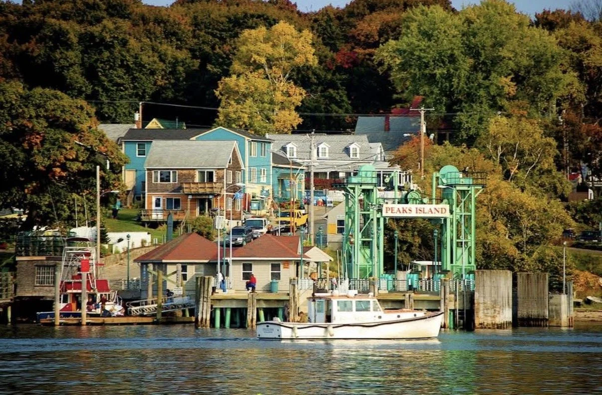 View of a waterfront community in Peaks Island, featuring colorful houses, a boat on the water, and a funicular incline with a sign reading 'PEAKS ISLAND'.