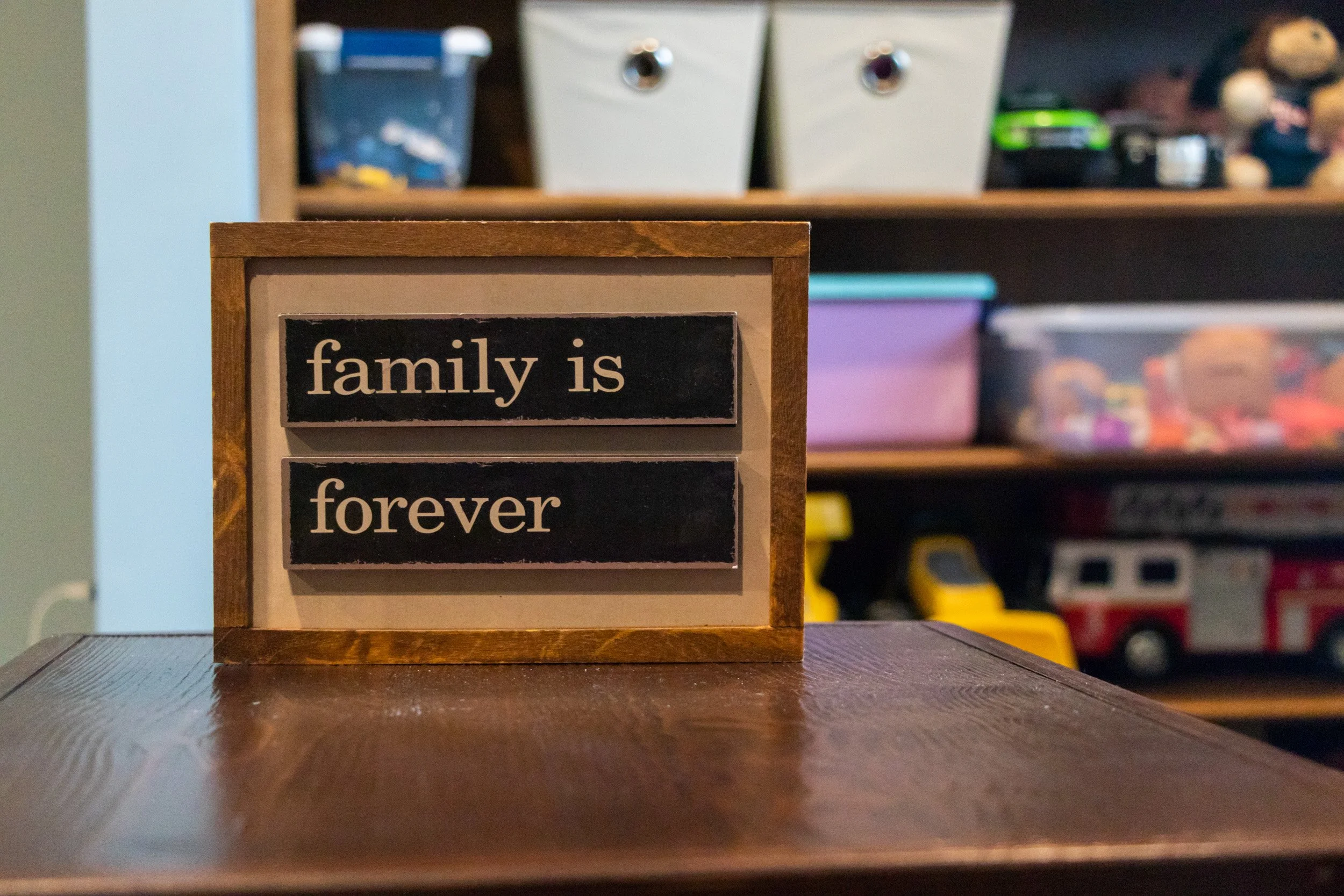A wooden block with the words 'family is forever' written on it, placed on a table in a room with shelves of toys and storage containers in the background.
