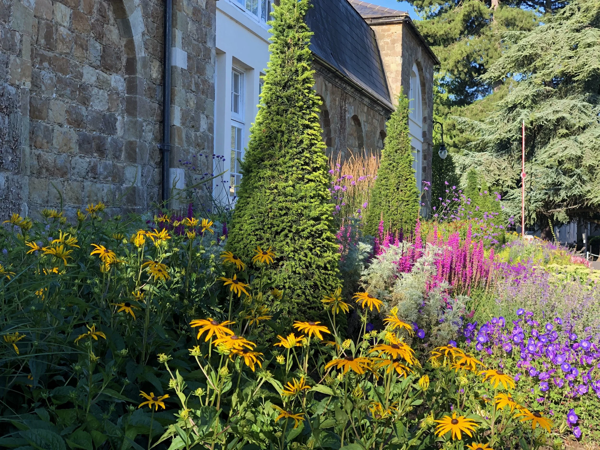 View of a garden with colorful flowers including yellow, purple, and pink, in front of a stone and white-walled building with windows and a sloped roof, surrounded by tall green trees.