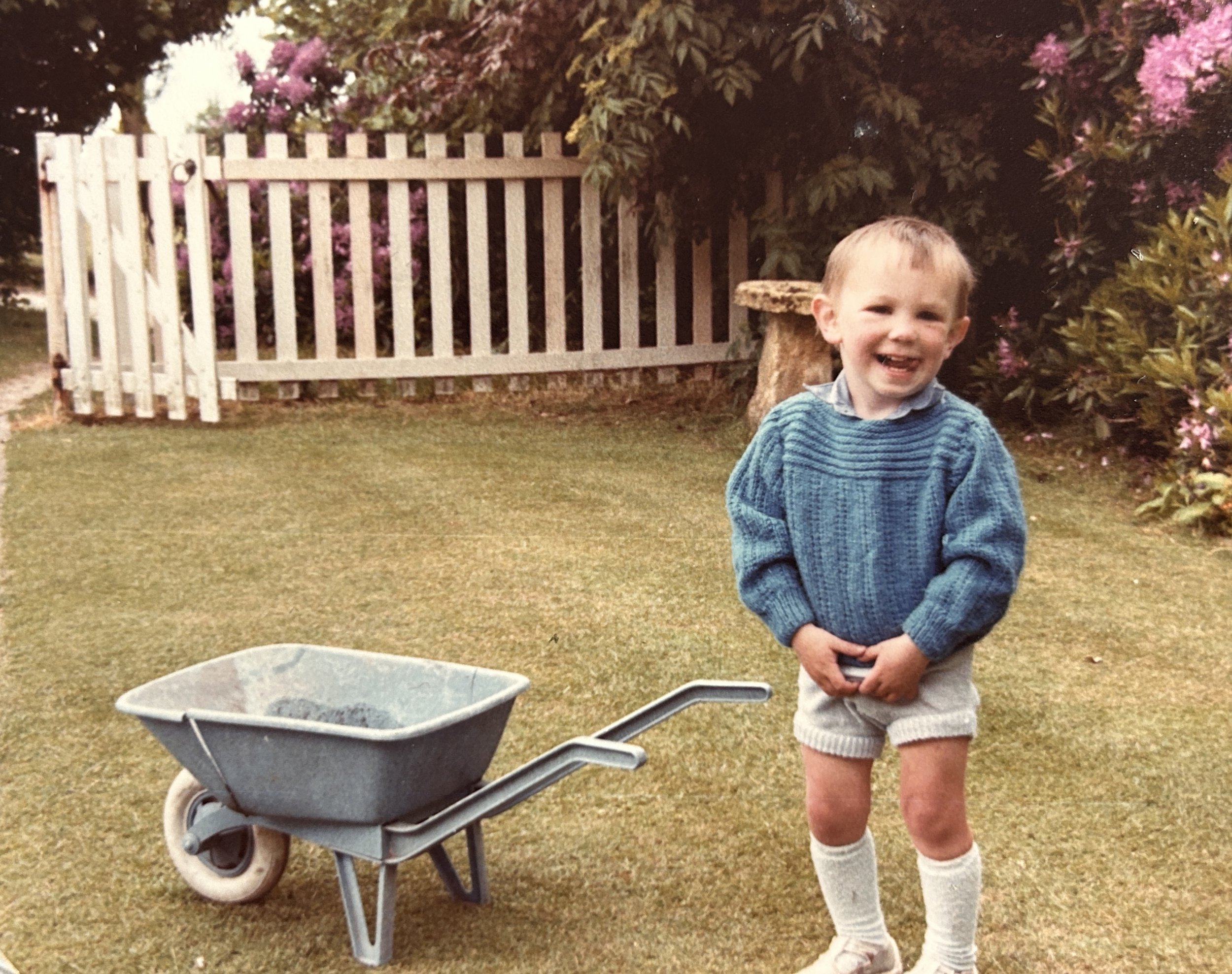 A young boy with short light hair, wearing a blue sweater, cream shorts, and knee-high socks, smiling and holding his hands in front of him outdoors in a garden with pink blooming bushes, next to a gray wheelbarrow filled with dirt.