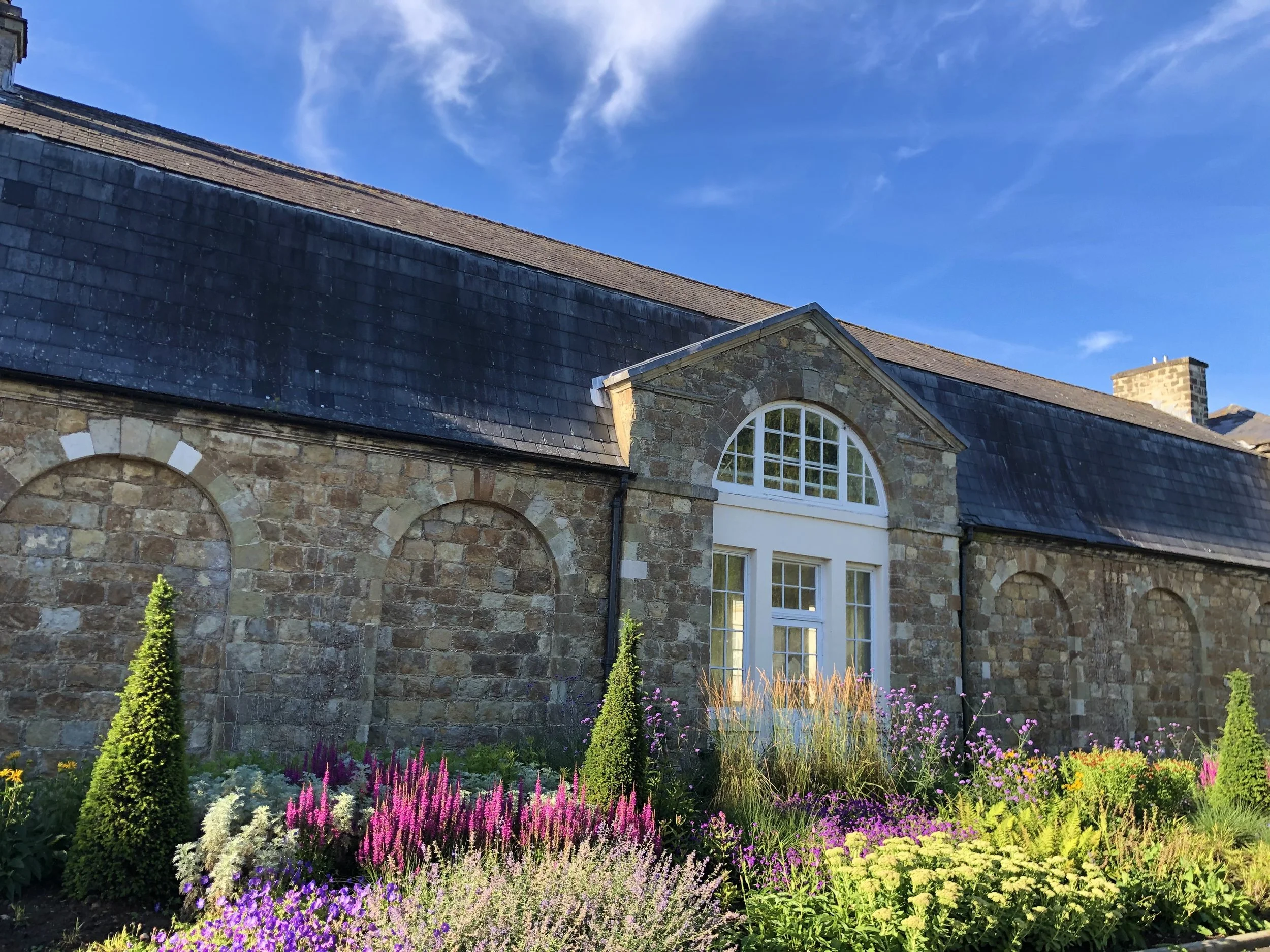 A stone house with a dormer window and slate roof, in a garden with colorful flowers and green shrubs, under a blue sky with some clouds.
