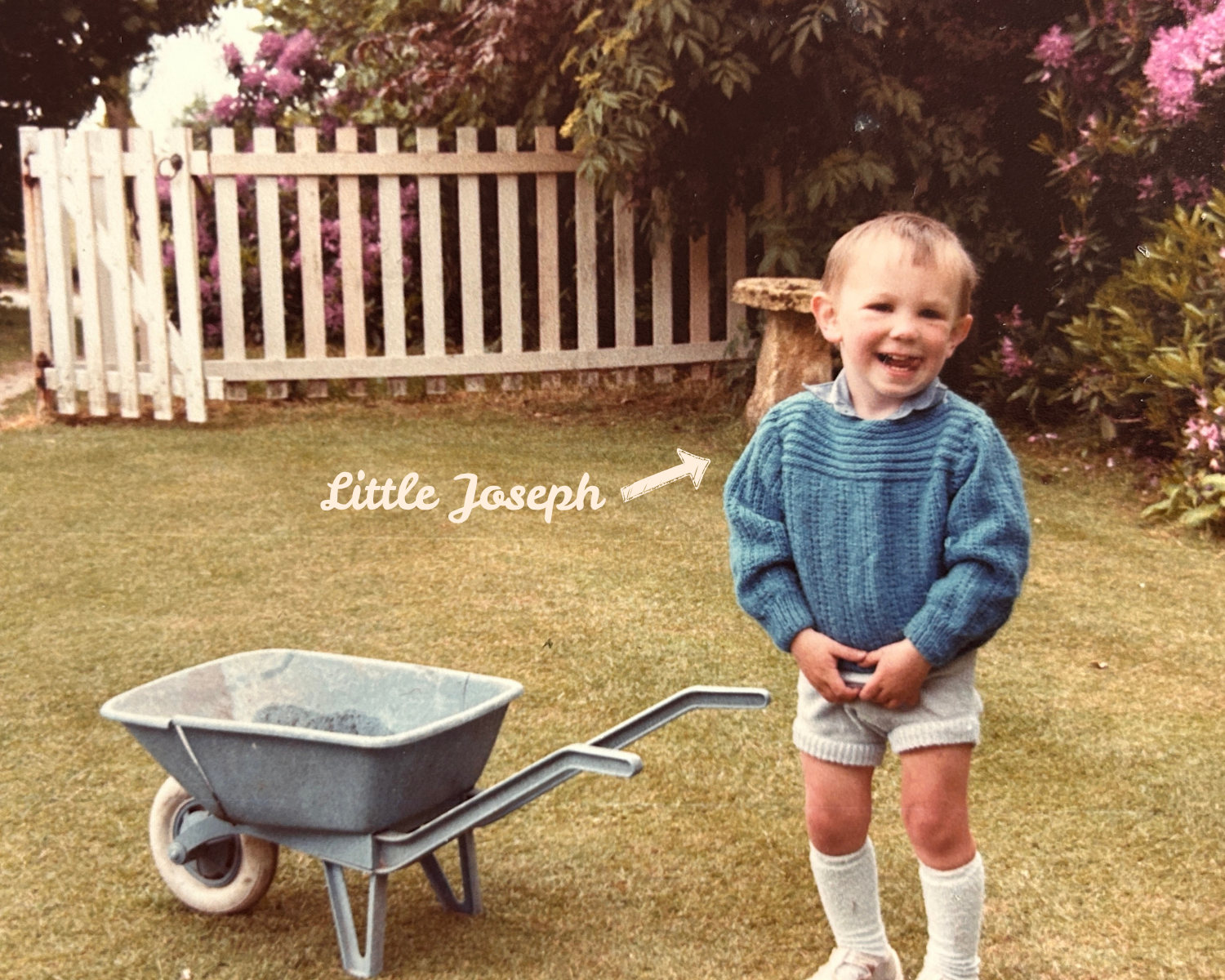 JP Gardens gardening expert at 4 in shorts standing in a garden, smiling, with a wheelbarrow nearby; background features a white picket fence, pink flowering bushes, and trees, and freshly cut grass.