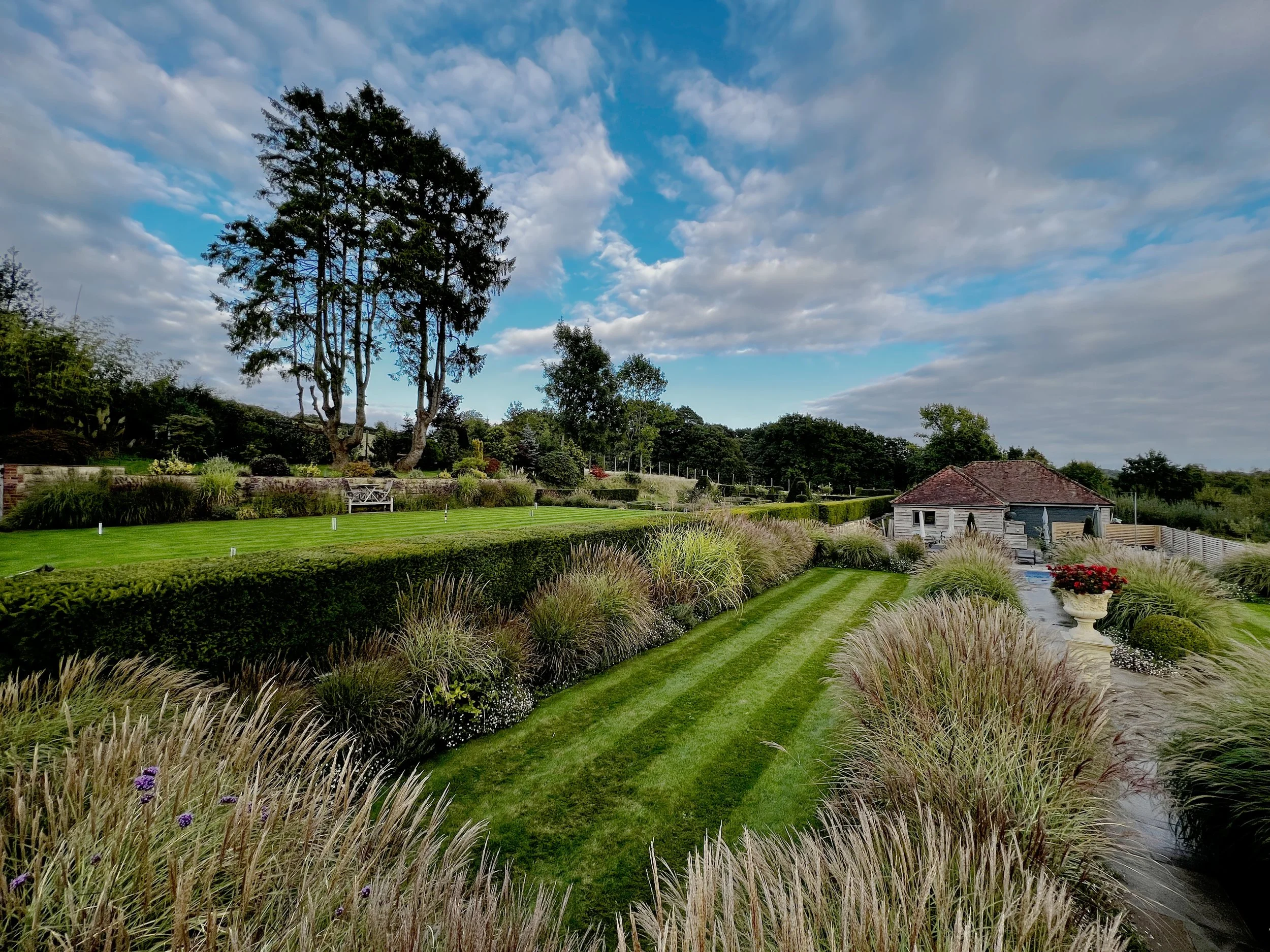 A landscaped garden with neatly trimmed striped grass, ornamental plants, and shrubs. There is a gravel path running through the garden, a land of many acres. Tall trees and a cloudy sky are in the background. Garden maintained by JP Gardens, Kent. 