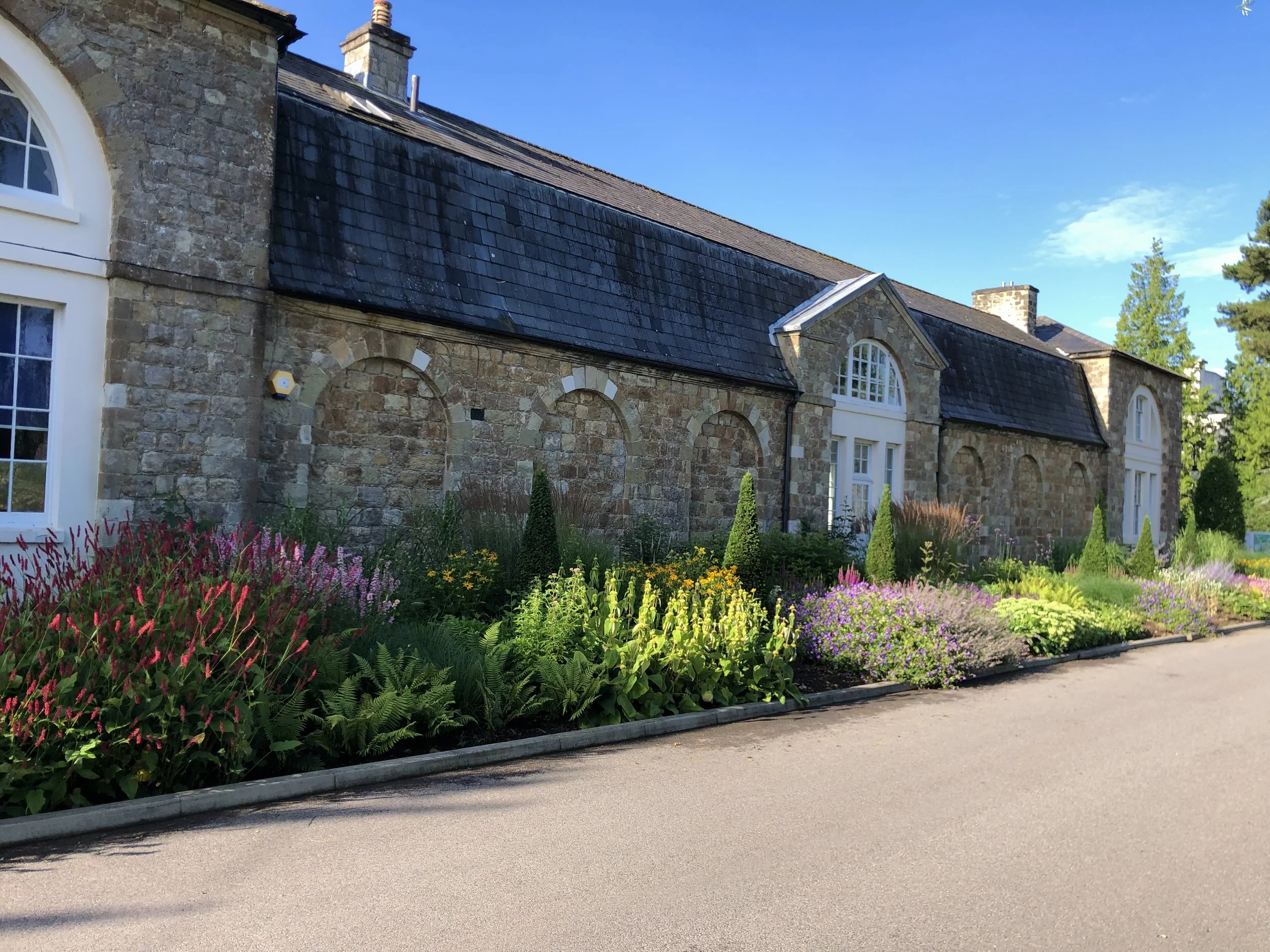 A stone and stucco house with arched windows, a slate roof, and a garden with colorful flowers and green shrubs, next to a paved road, under a clear blue sky.