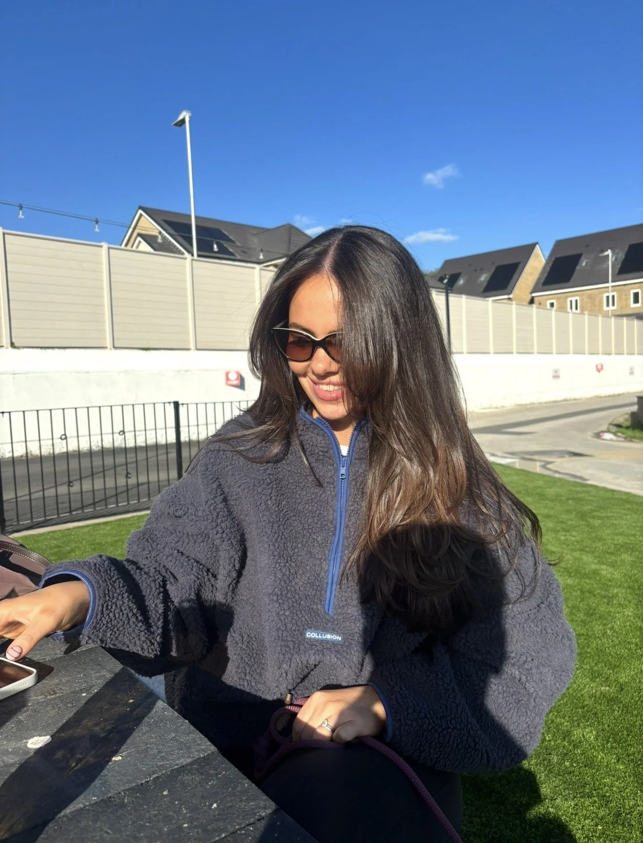 Woman wearing sunglasses and a dark fleece jacket seated outdoors at a picnic table on a sunny day