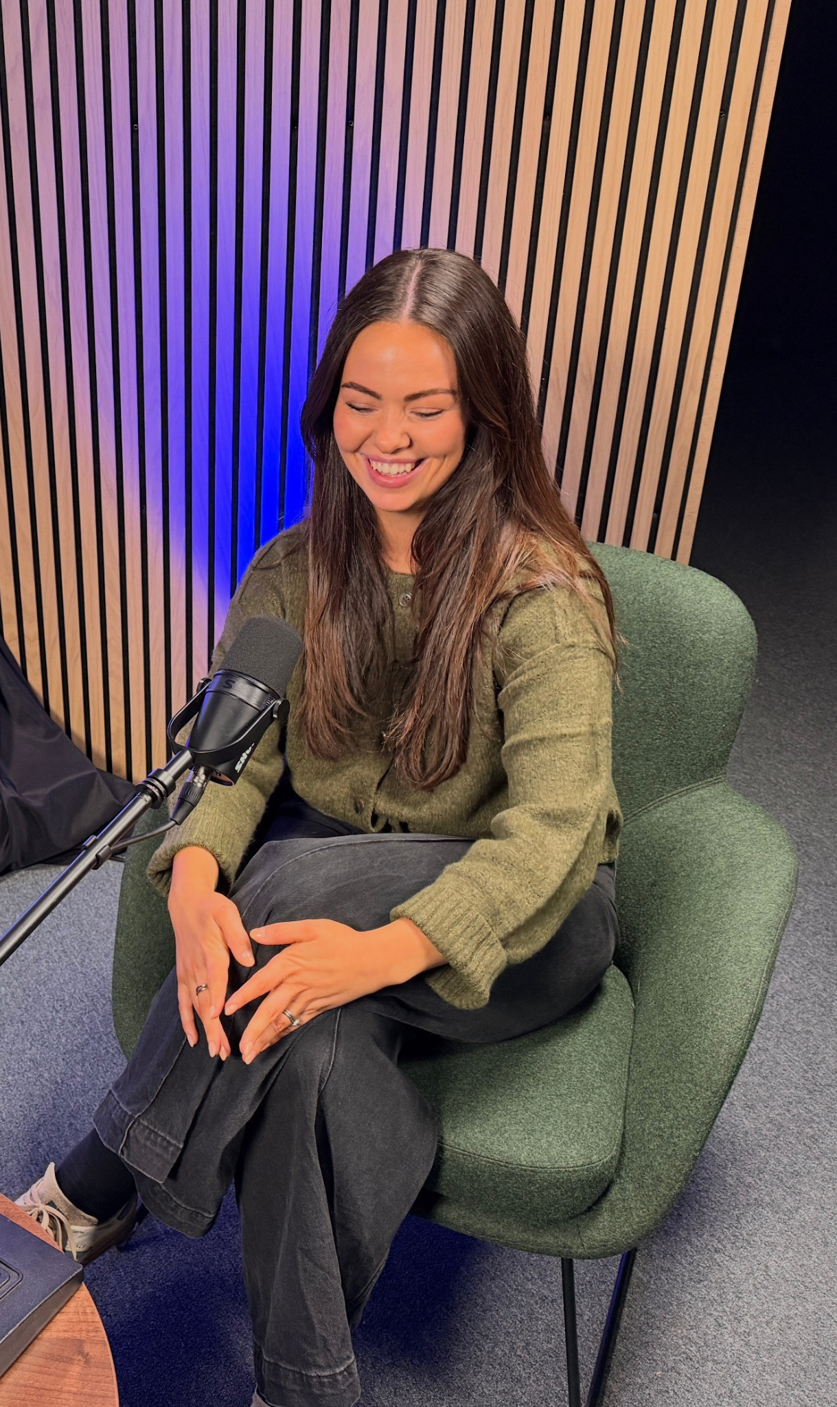 Smiling woman seated in a green armchair beside a podcast microphone in a recording studio